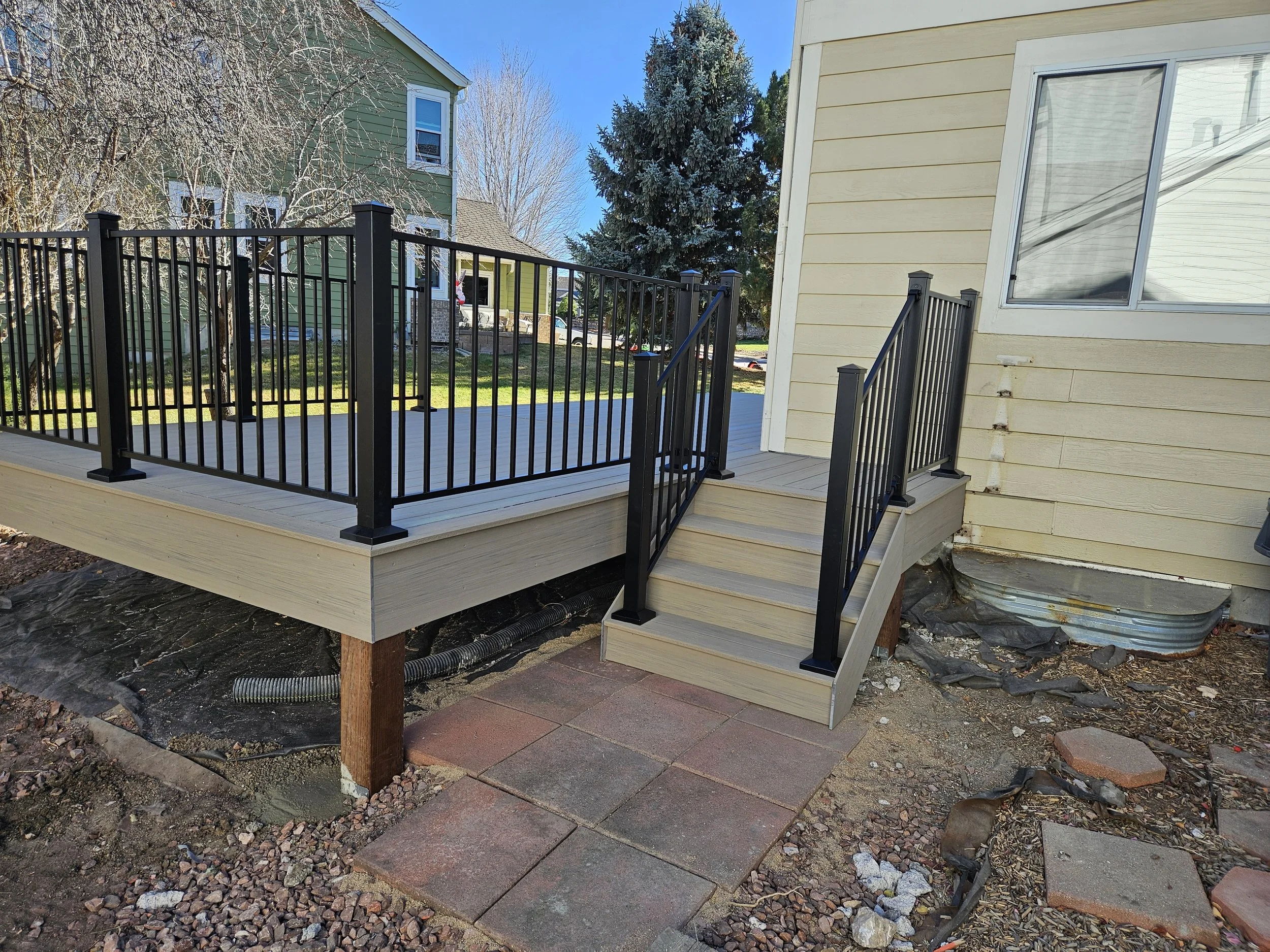 Newly installed wooden deck with black metal railing and stairs attached to a yellow house with a large window, in a backyard with trees and neighboring houses.
