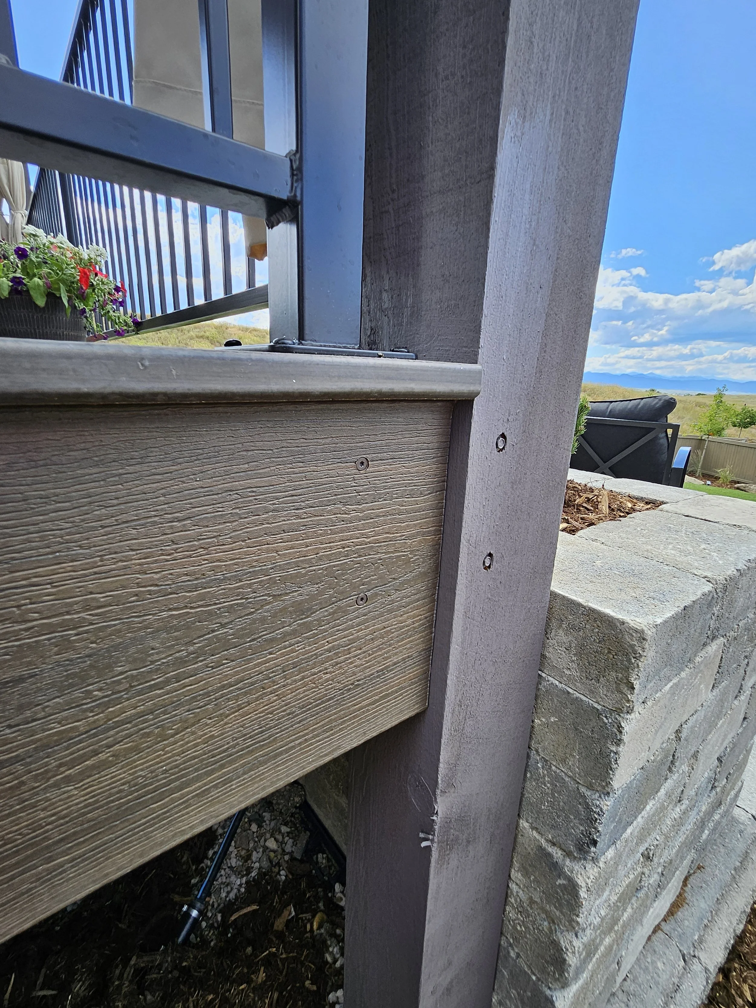 Close-up of a deck post with a wooden rail, a metal railing, a gray brick wall, and a concrete patio with outdoor furniture and a landscape with blue sky and clouds in the background.