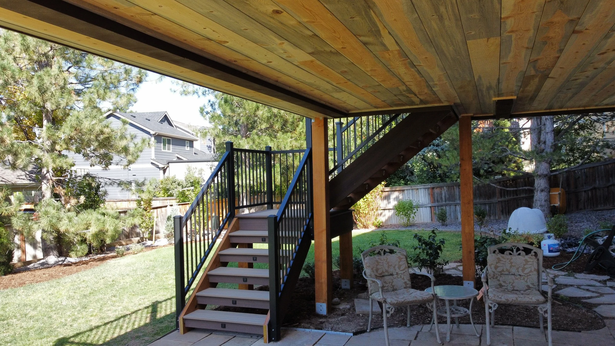 Backyard patio area with stairs leading to a covered deck, surrounded by trees, yard chairs, and gardening tools.