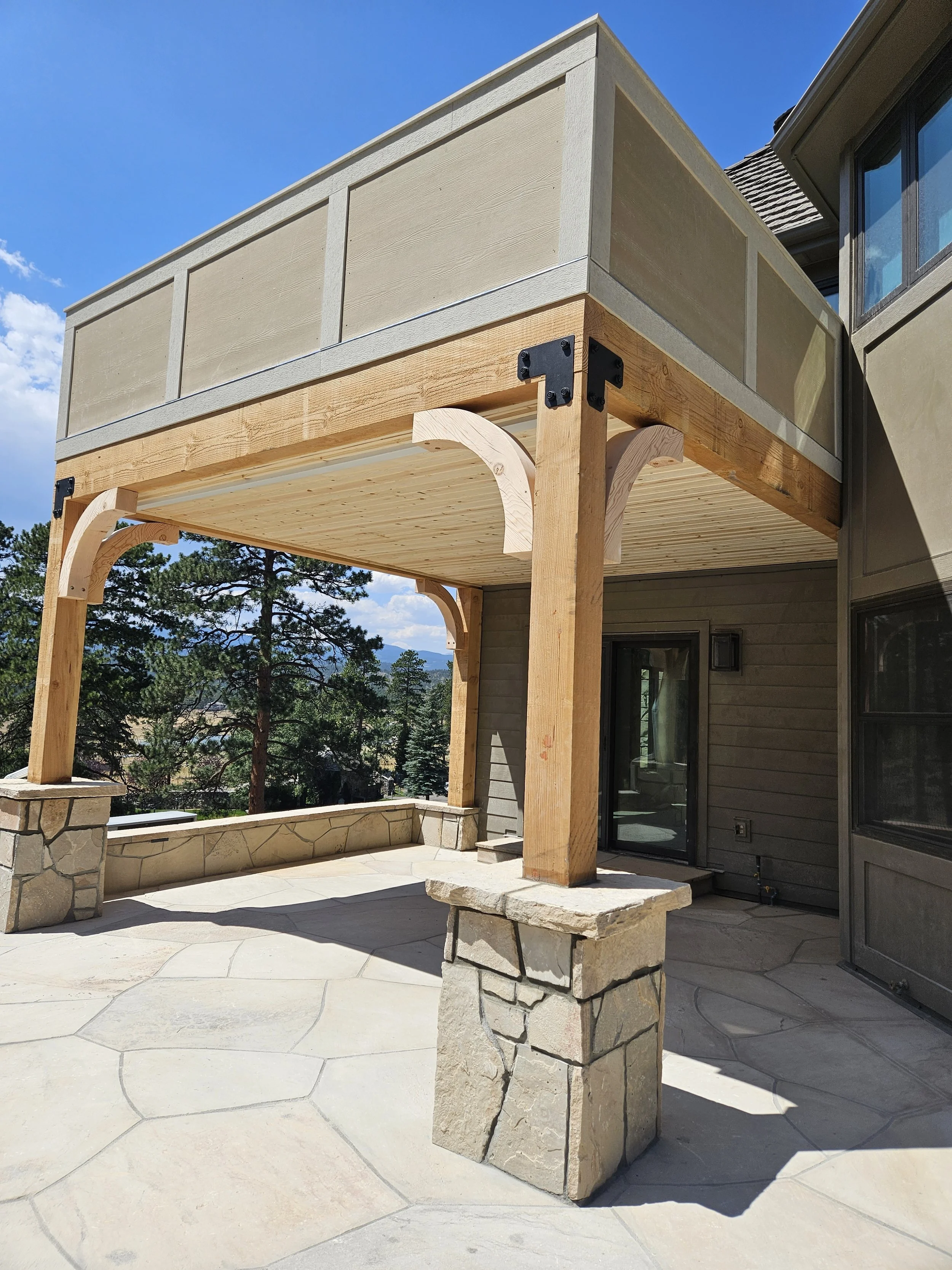 A partially constructed wooden patio with stone pillars, underneath a balcony with beige railing, overlooking a mountain and trees landscape.