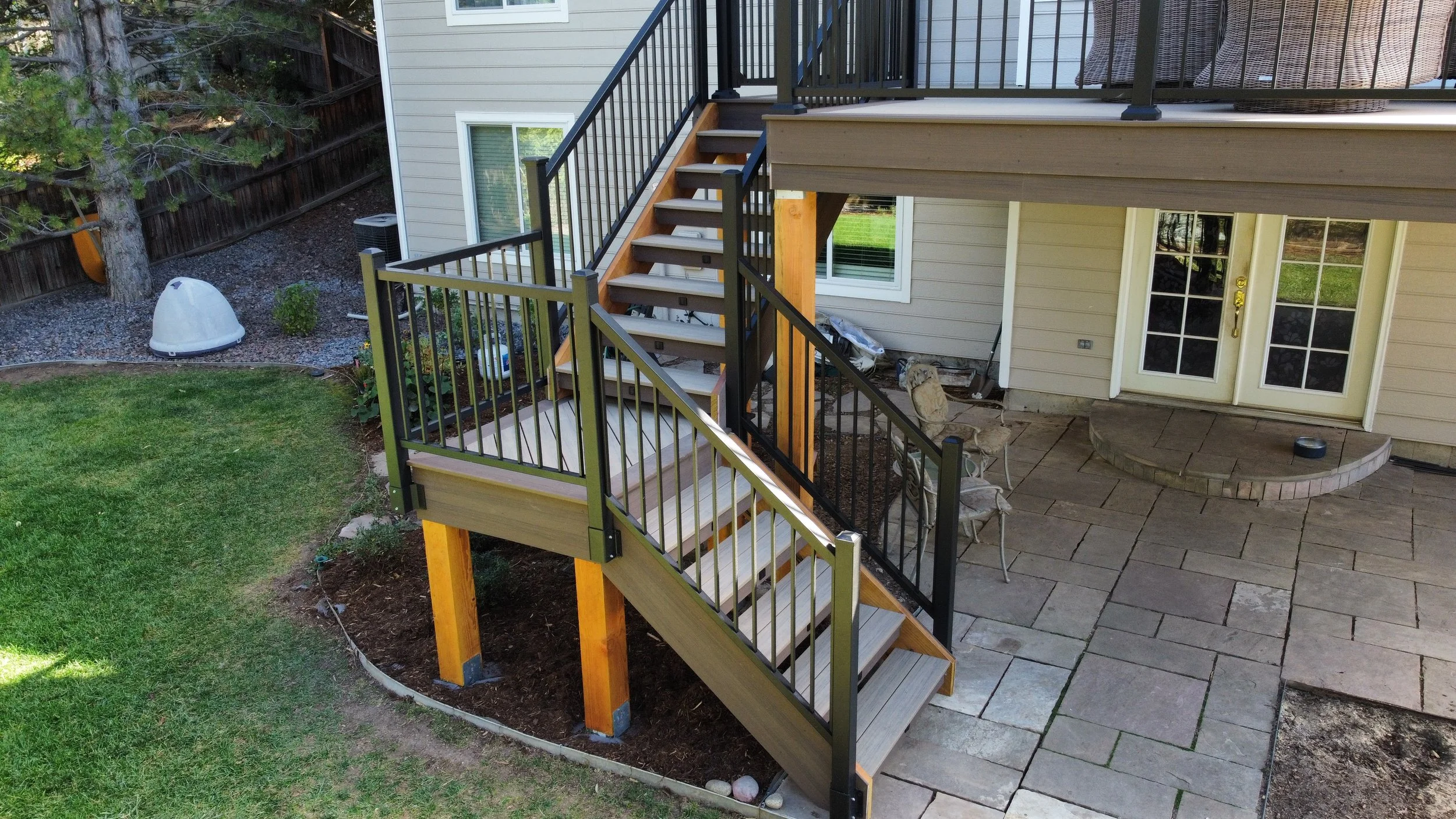 View of a backyard with a newly built staircase leading from an elevated deck to the ground, with a patio area, grass, and trees in the background.