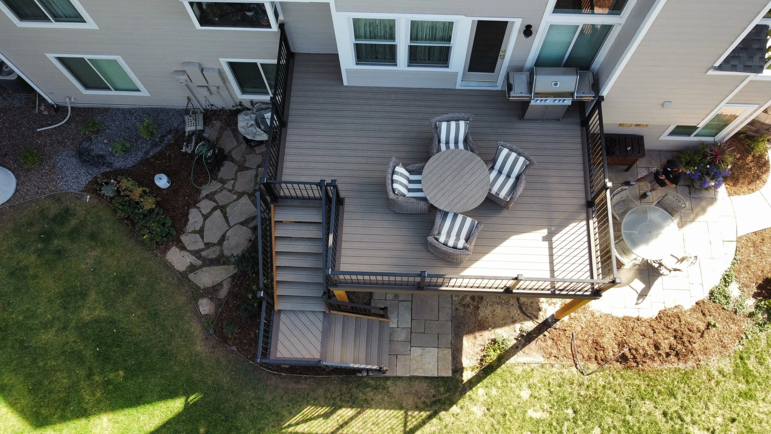 Top-down view of a residential backyard deck with a round table and four striped cushioned chairs, attached to a house with sliding glass doors and an outdoor grill, bordered by a garden bed, a grassy lawn, and a paved patio area where a person is si