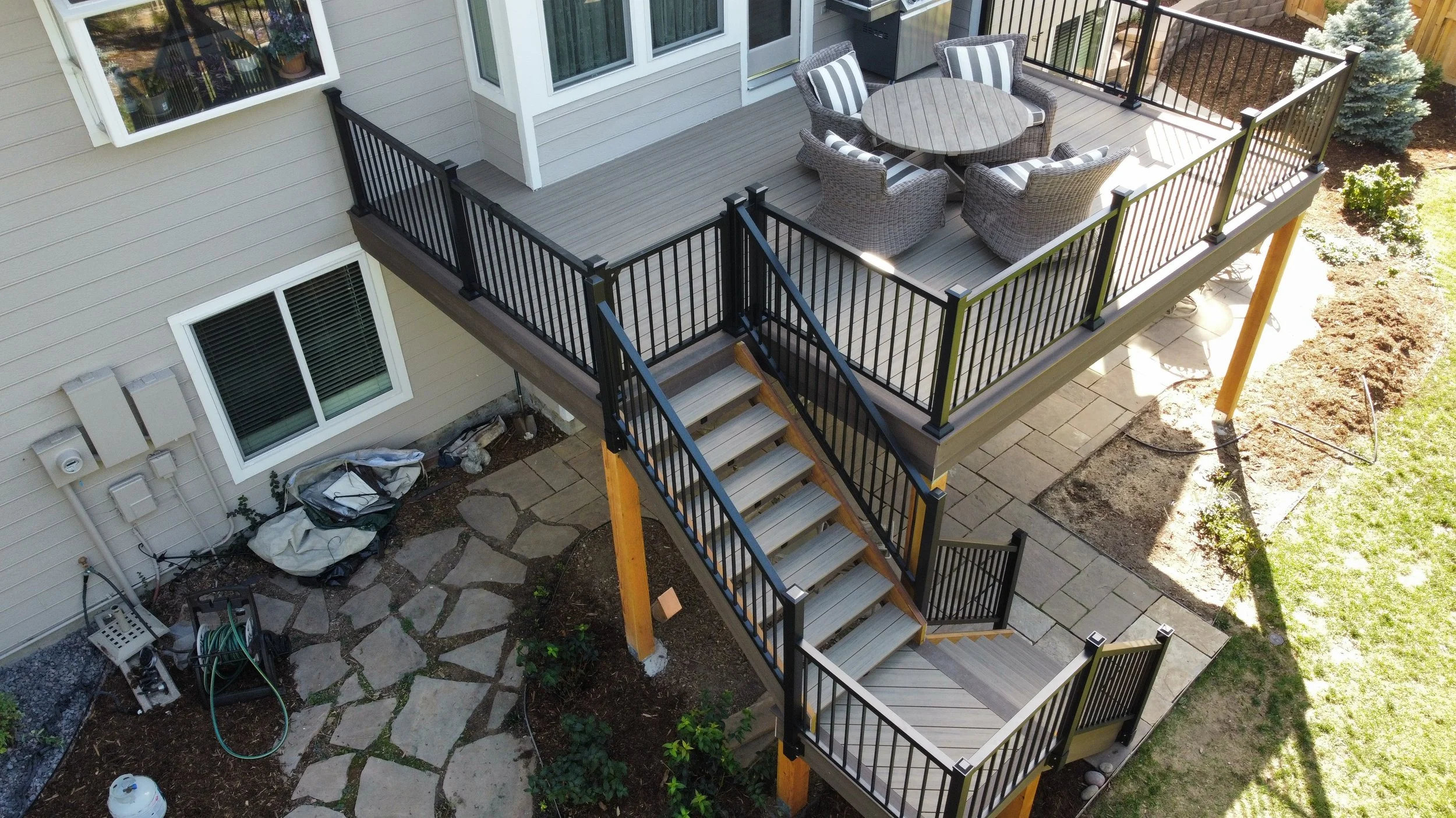 An aerial view of a two-story house backyard featuring a wooden deck with outdoor furniture, black metal railing, and a staircase leading down to a stone patio with gardening supplies and a small garden area.