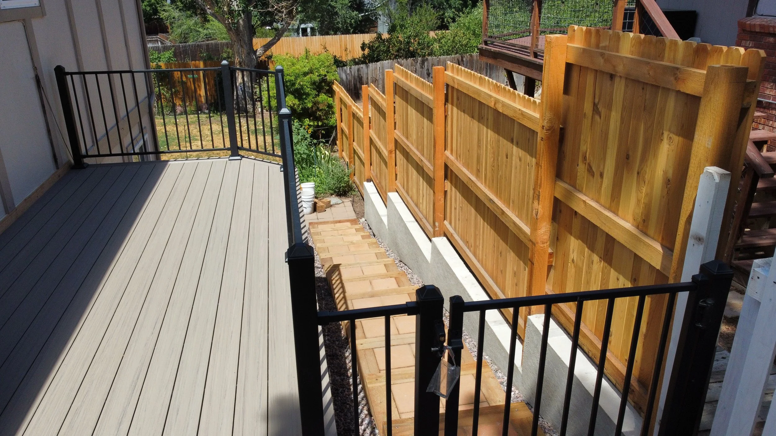 A backyard with a newly installed wooden fence on the right side, a black metal railing on the left, a deck with light-colored wood planks, and a large tree with green foliage in the background by Mile High Decks Specializing in Decks and Outdoor