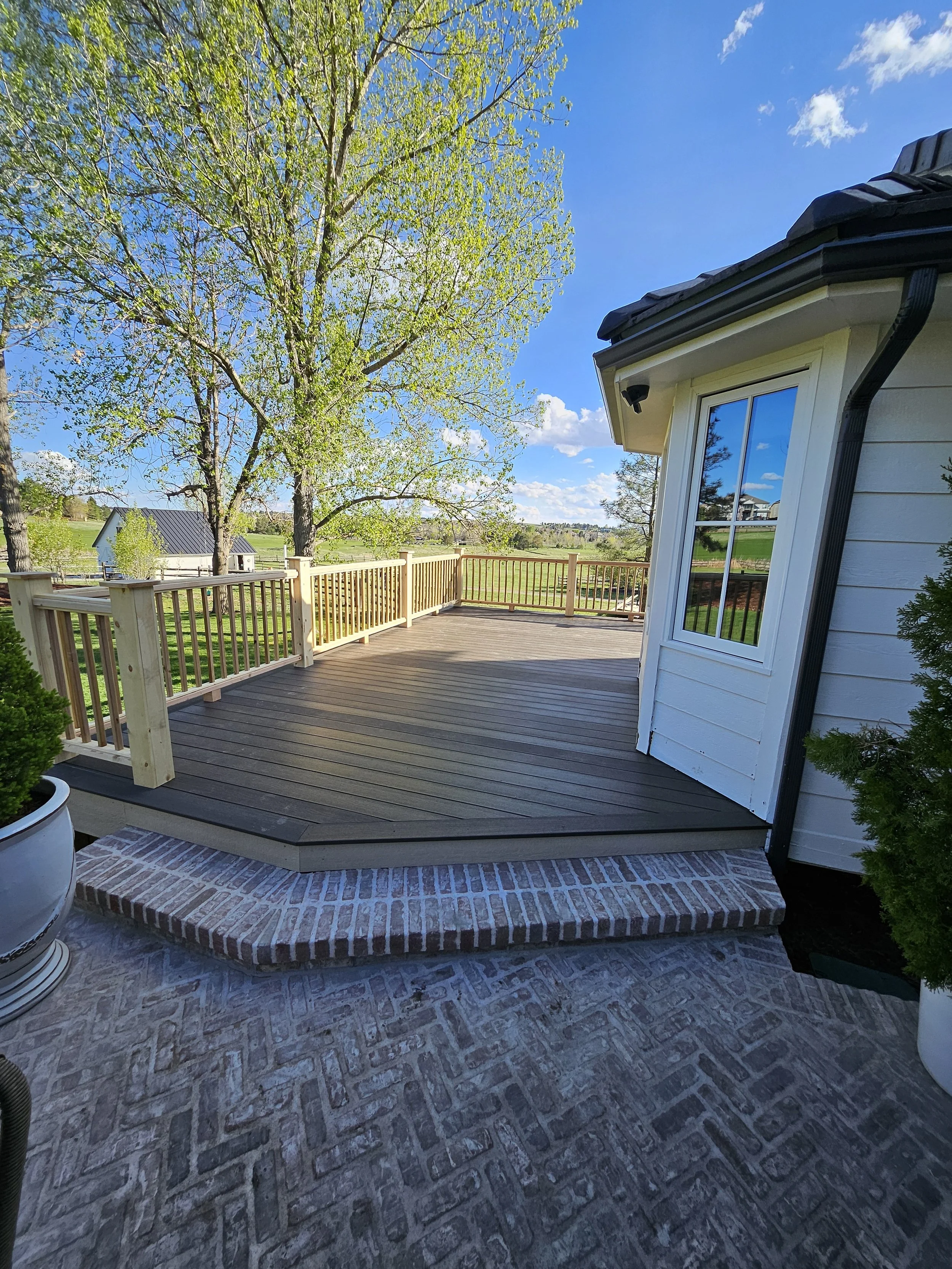 A new wooden deck attached to a house with white siding, a large window, and brick steps leading up to the deck. The deck overlooks a green field with trees and a small white building in the distance on a sunny day.