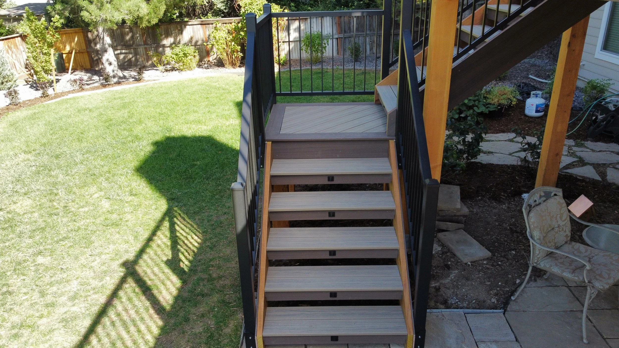 Backyard view of a wooden staircase with black metal railings leading up to a deck. The yard has a well-kept lawn, a garden with plants and trees, and garden path stones. There is an outdoor chair with patterned cushion on the patio.