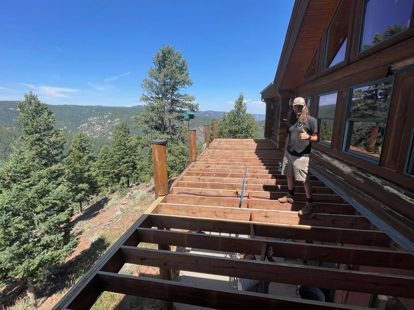 A woman in a beige hat and black shirt stands on a wooden deck under construction, giving a thumbs-up, with a scenic mountain landscape and trees in the background.