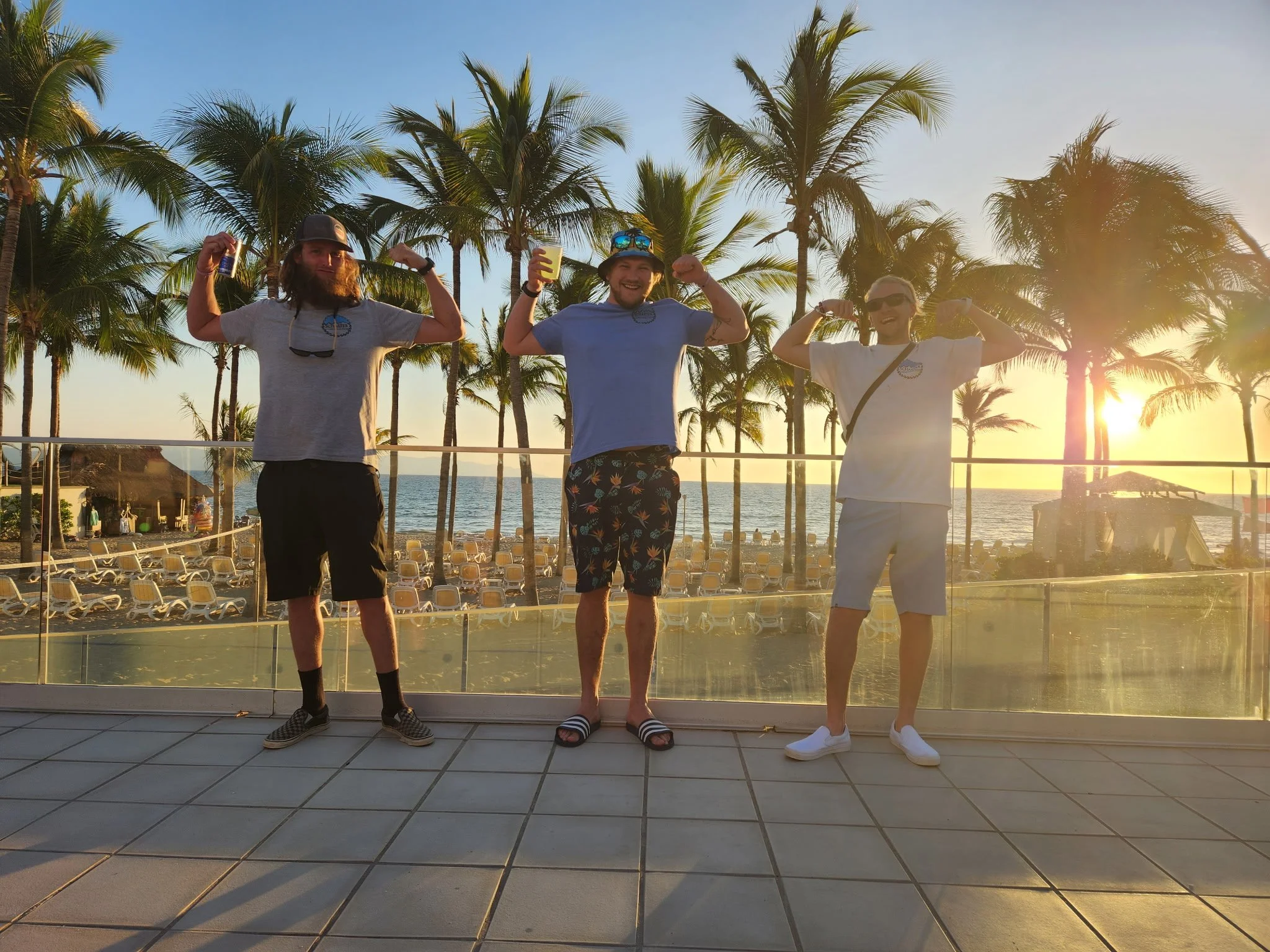 Three men standing on a balcony with palm trees and the ocean in the background, flexing their biceps during sunset, with beach chairs and a hut visible below.