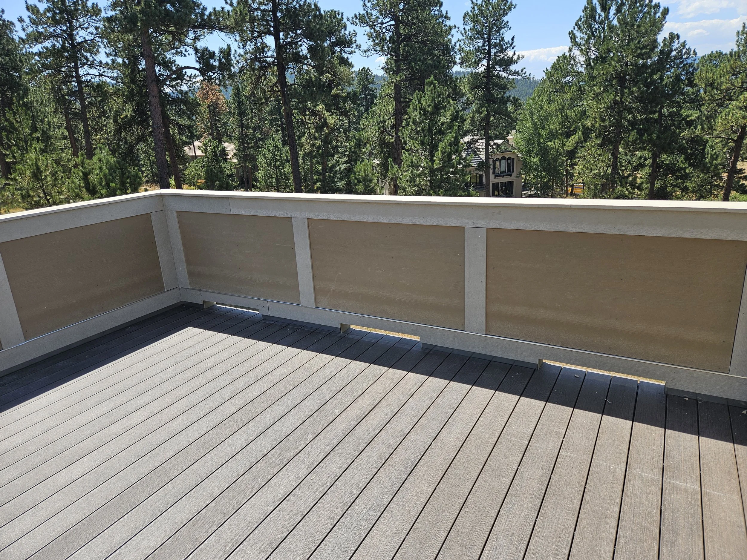 View of a balcony with a wooden floor and beige railing, overlooking a forested area with trees and a distant building under a partly cloudy sky.