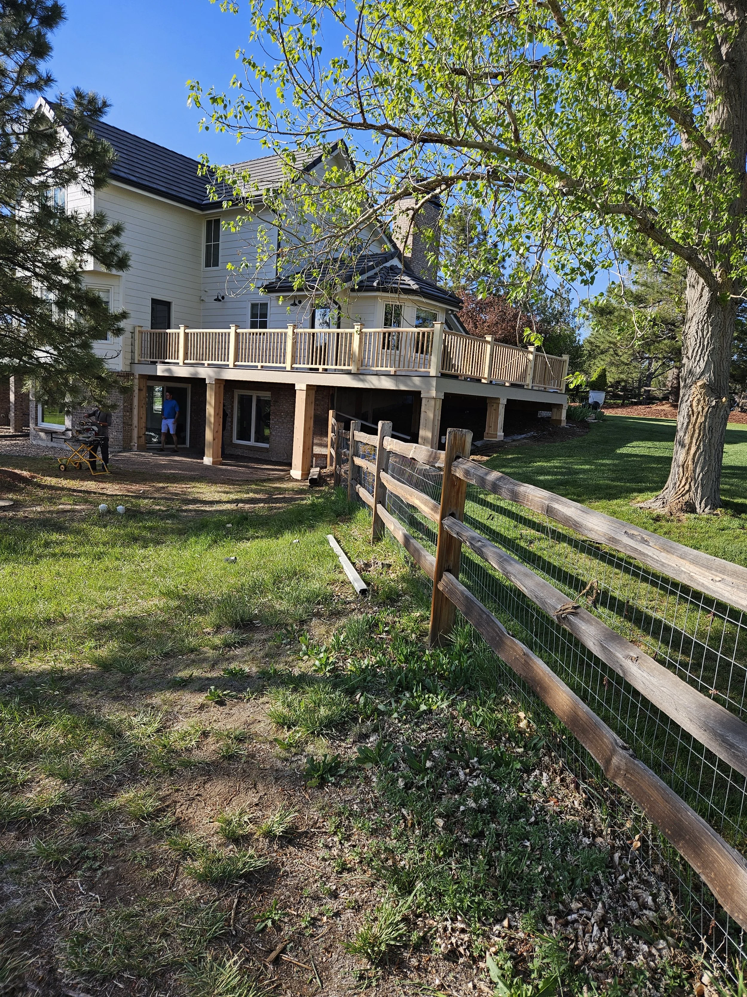 A two-story house with a newly built wooden deck and railing, surrounded by green trees and grass, with a wooden fence in the foreground and a clear blue sky overhead.