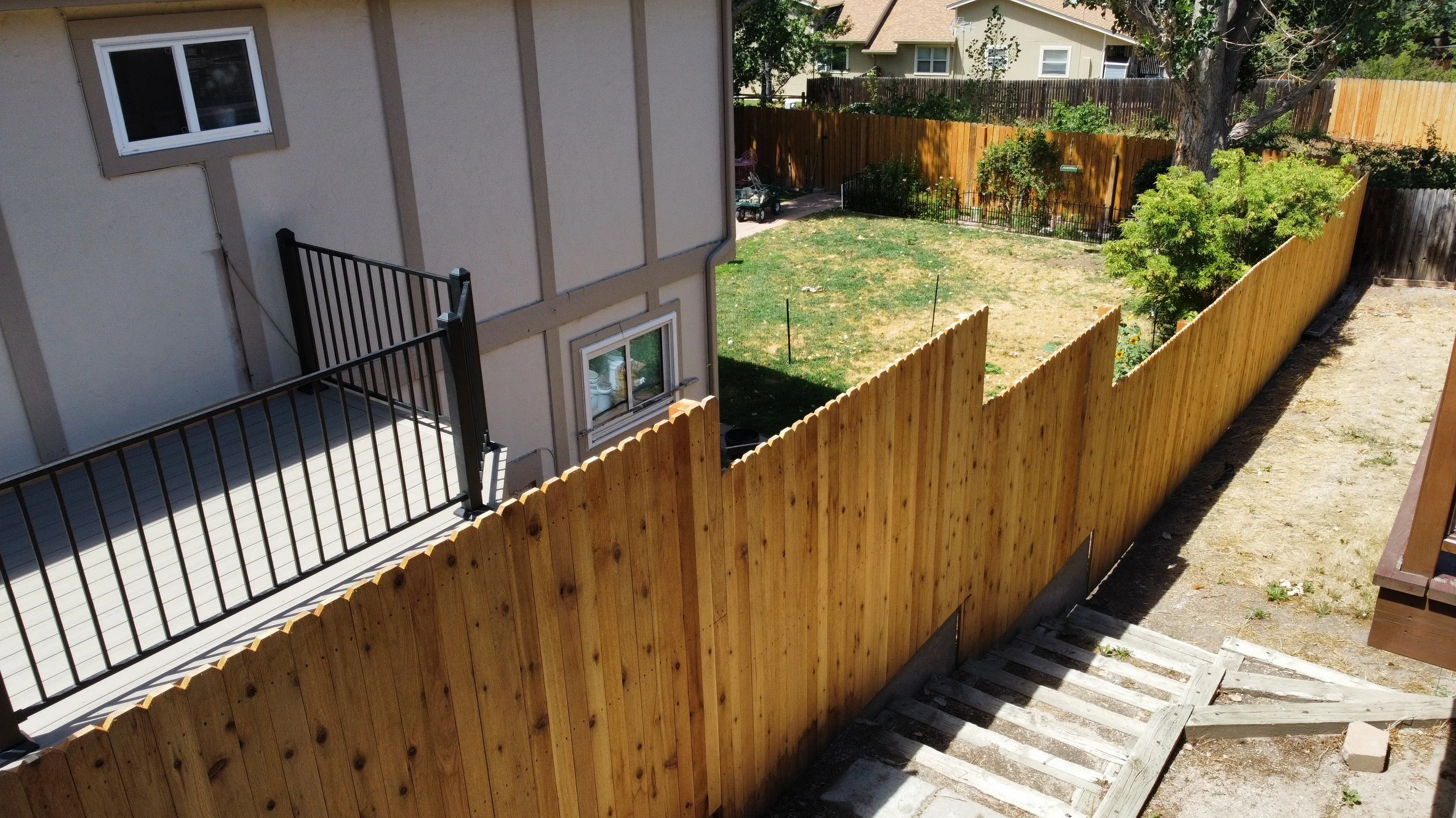 View of a backyard with a wooden fence, a small lawn area, a tree, and neighboring houses in the background. Part of a house's exterior wall and a balcony with a black railing are visible in the foreground. Mile High Decks Specializing in Decks 