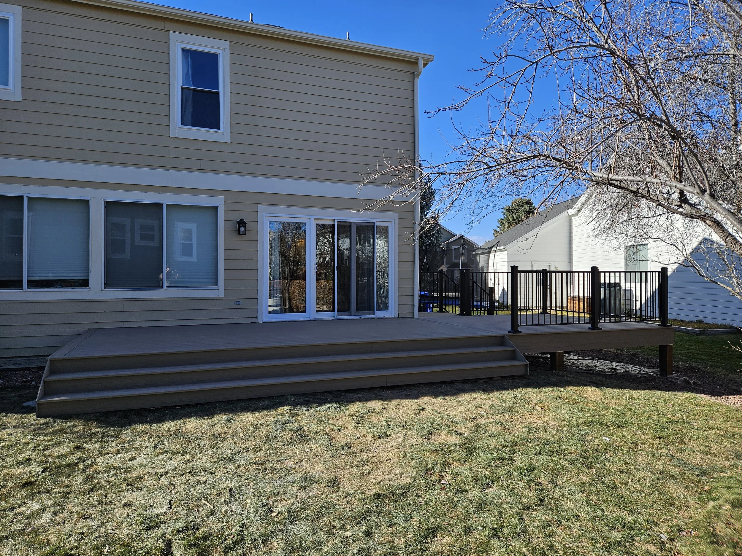 Backyard with a beige house, sliding glass door, and wooden deck with stairs. White fence visible in neighboring yard. Leafless tree in foreground on the right. Clear blue sky.