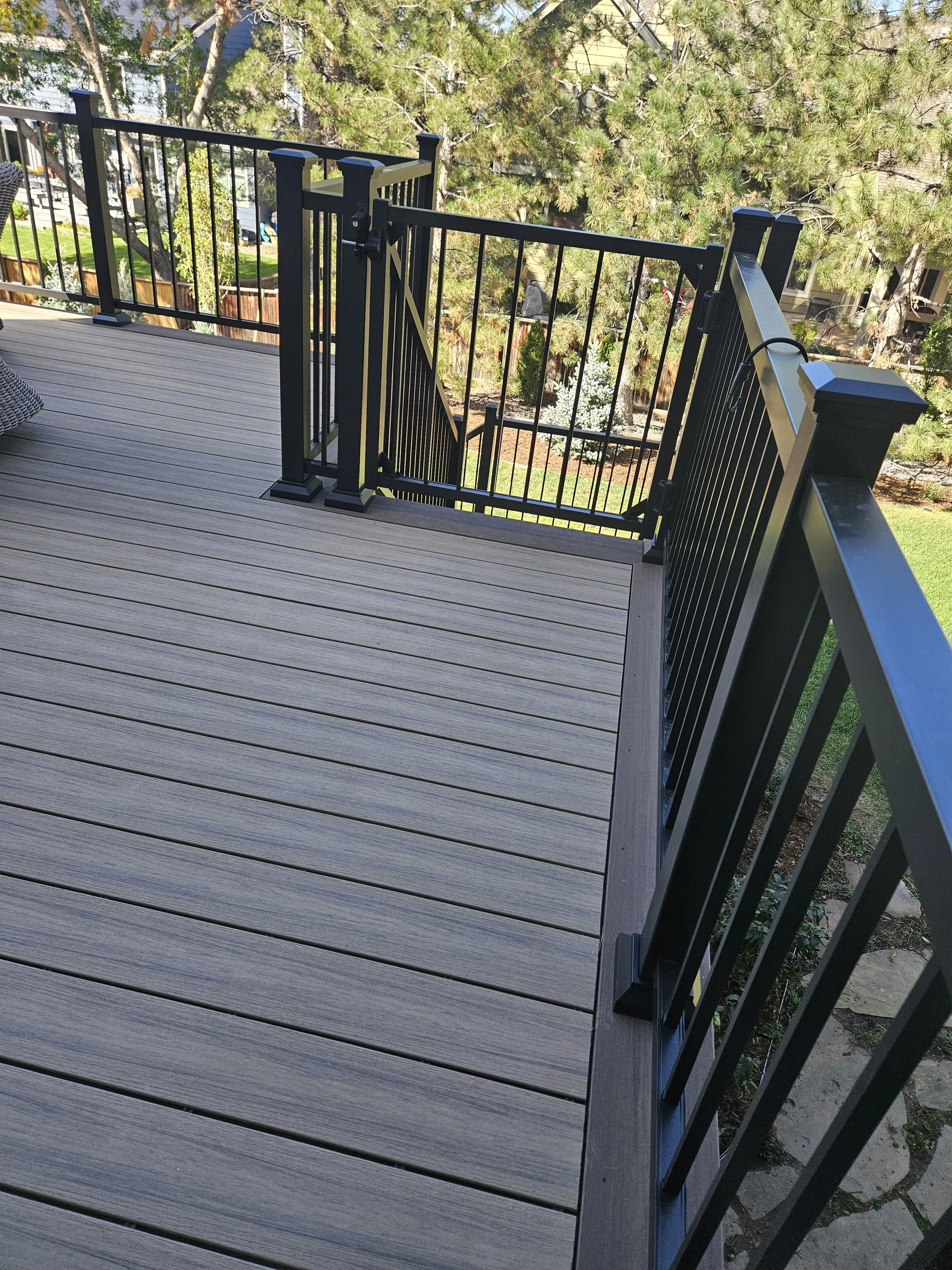 View of a backyard deck with black metal railing and staircase, overlooking a garden with trees and grass.