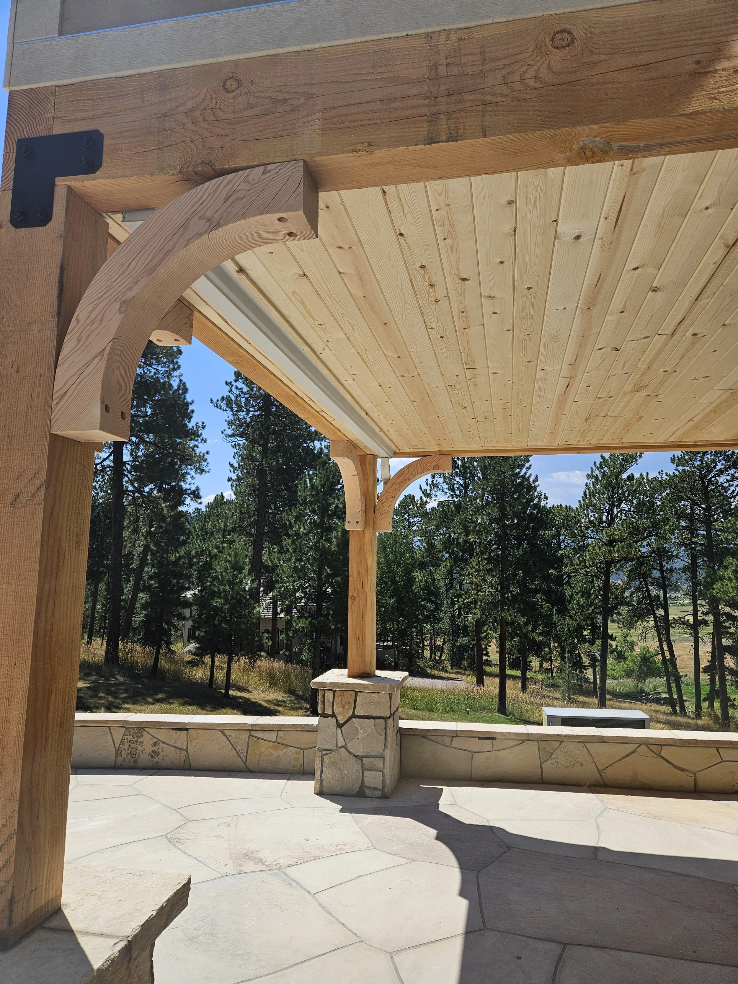 Close-up view of a wooden patio ceiling with decorative corbels, supported by a stone pillar, overlooking a forested landscape on a sunny day.