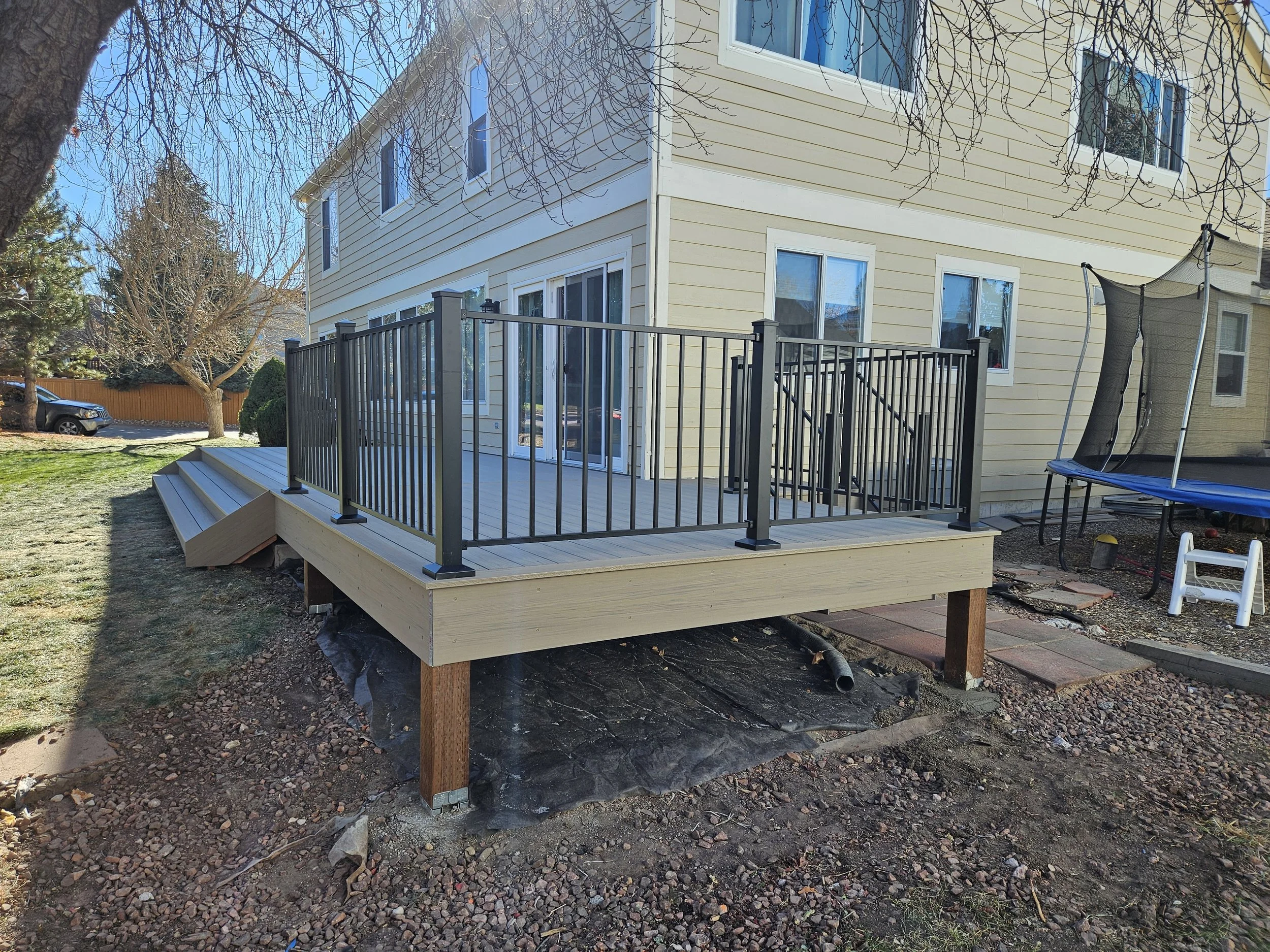 A newly built wooden deck attached to the back of a yellow house with white trim and large glass sliding doors, surrounded by a black metal railing, with a small staircase leading down to the yard.