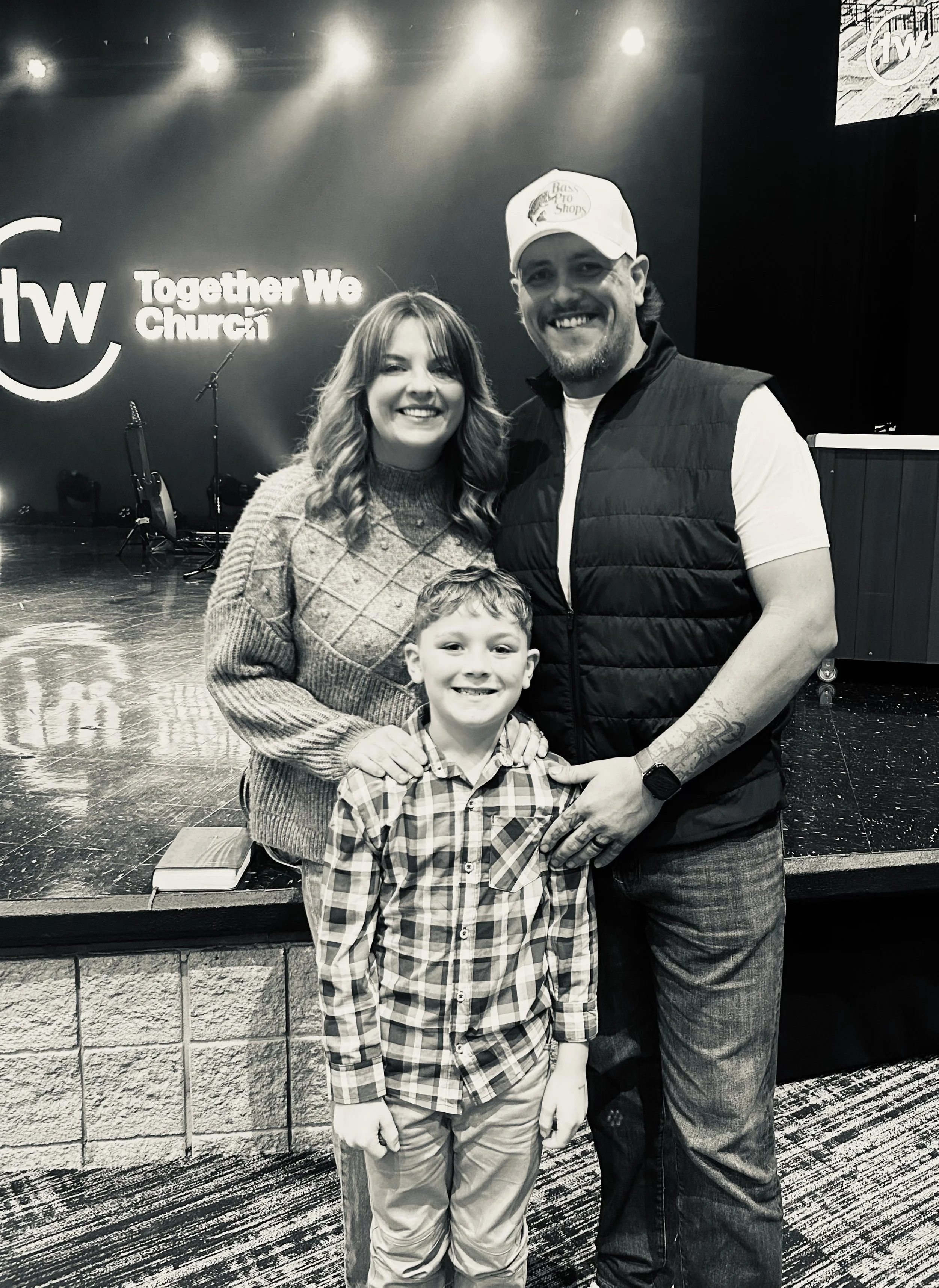A family of three smiling in front of a stage at a church event, with musical instruments and a sign reading 'Together We Church' in the background.