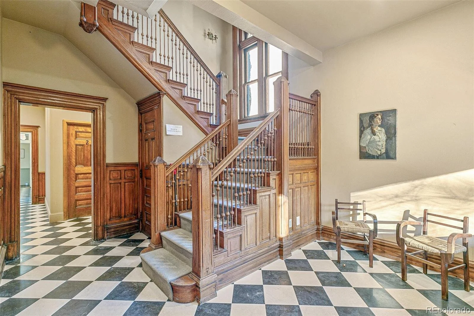 Interior view of a historic building with black and white checkered tile floor, wooden staircase, and vintage chairs. A portrait hangs on the wall opposite the staircase.