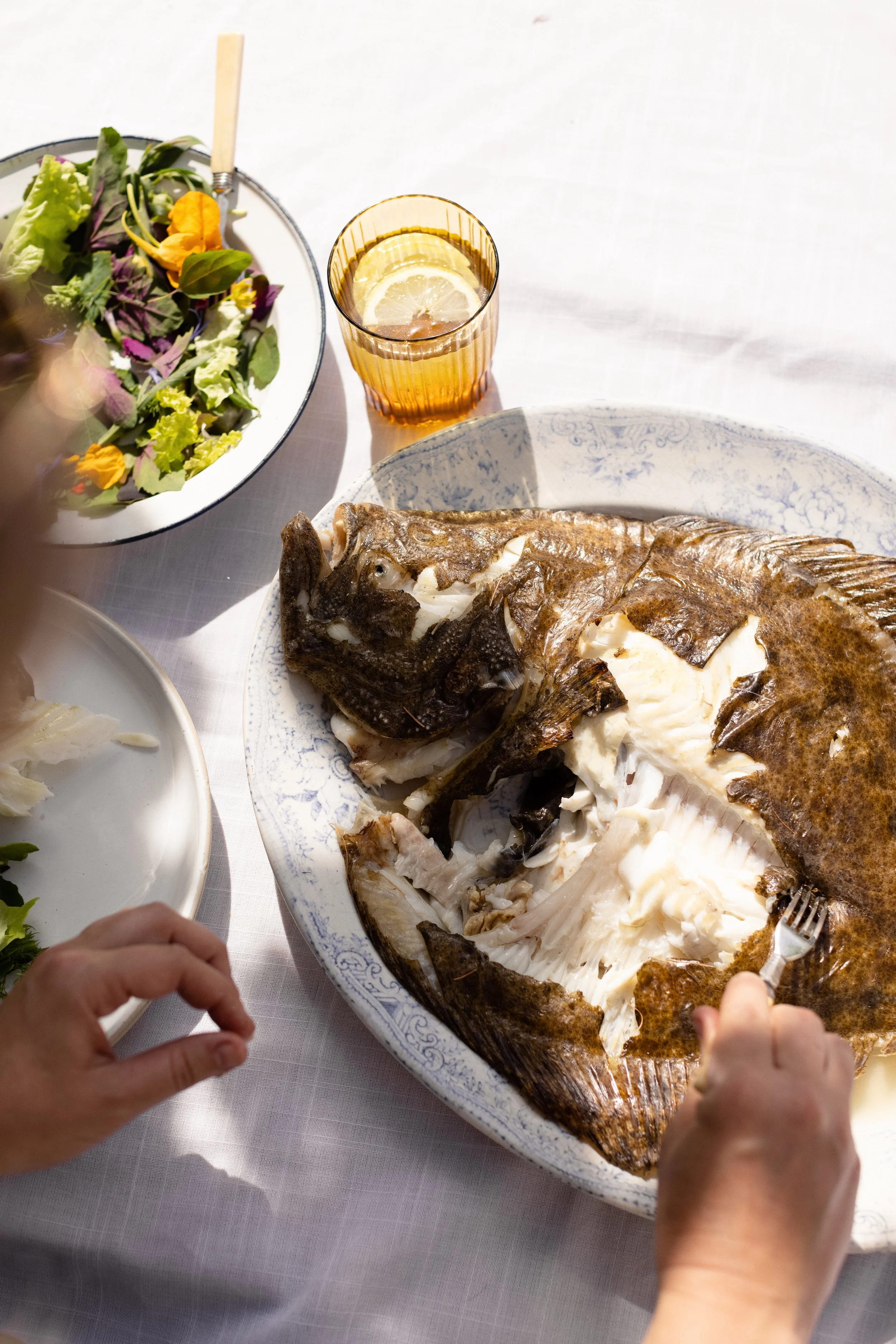 A person cutting into a cooked fish on a decorative plate at a dinner table with salad and a glass of lemon water.