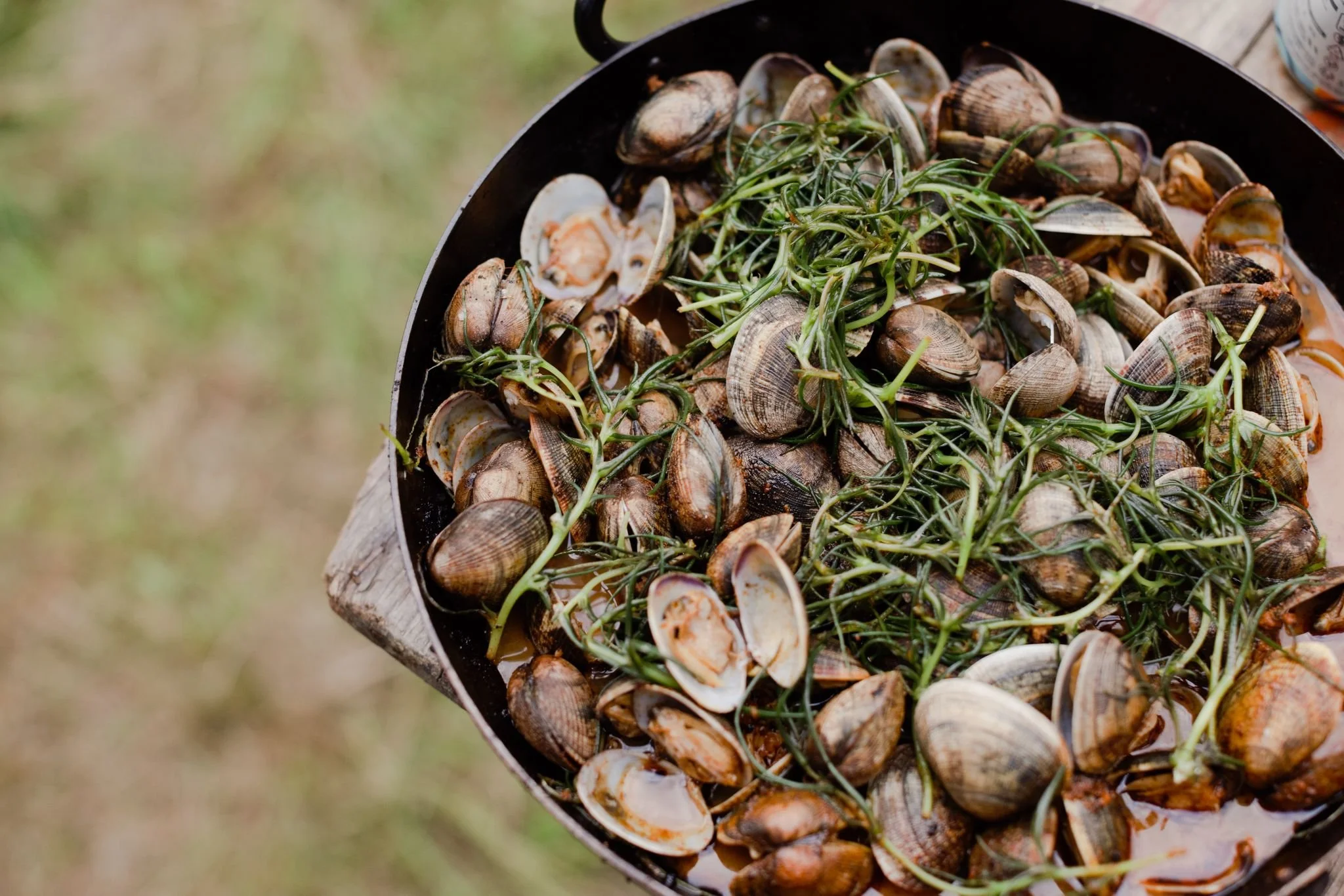 A black skillet filled with cooked clams and garnished with fresh green herbs, outdoors on a wooden surface.