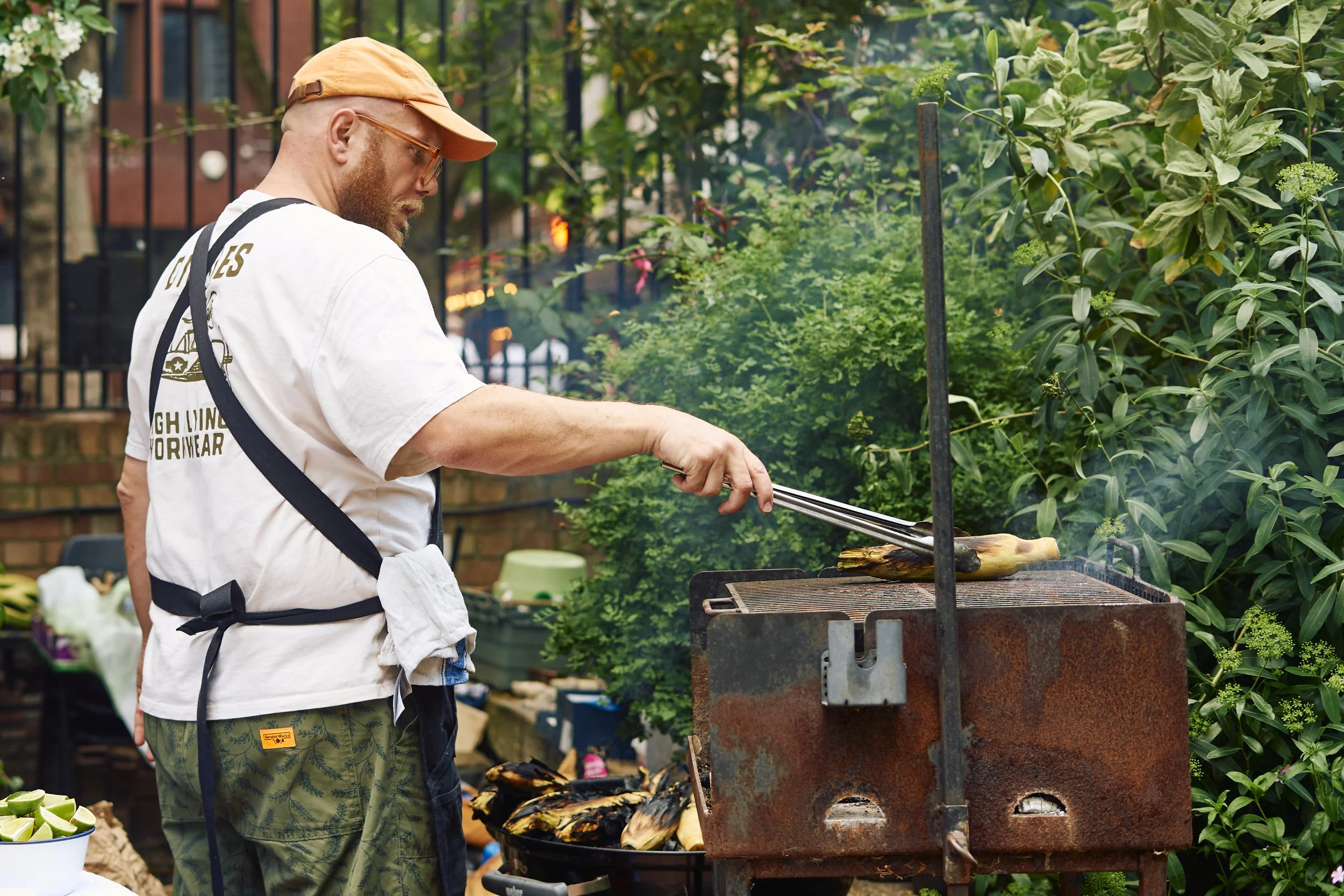 A man with a beard wearing an orange cap, sunglasses, white t-shirt, and camouflage pants is grilling corn on a barbecue grill outdoors. He is using tongs to turn the corn. There are lush green bushes and trees in the background.