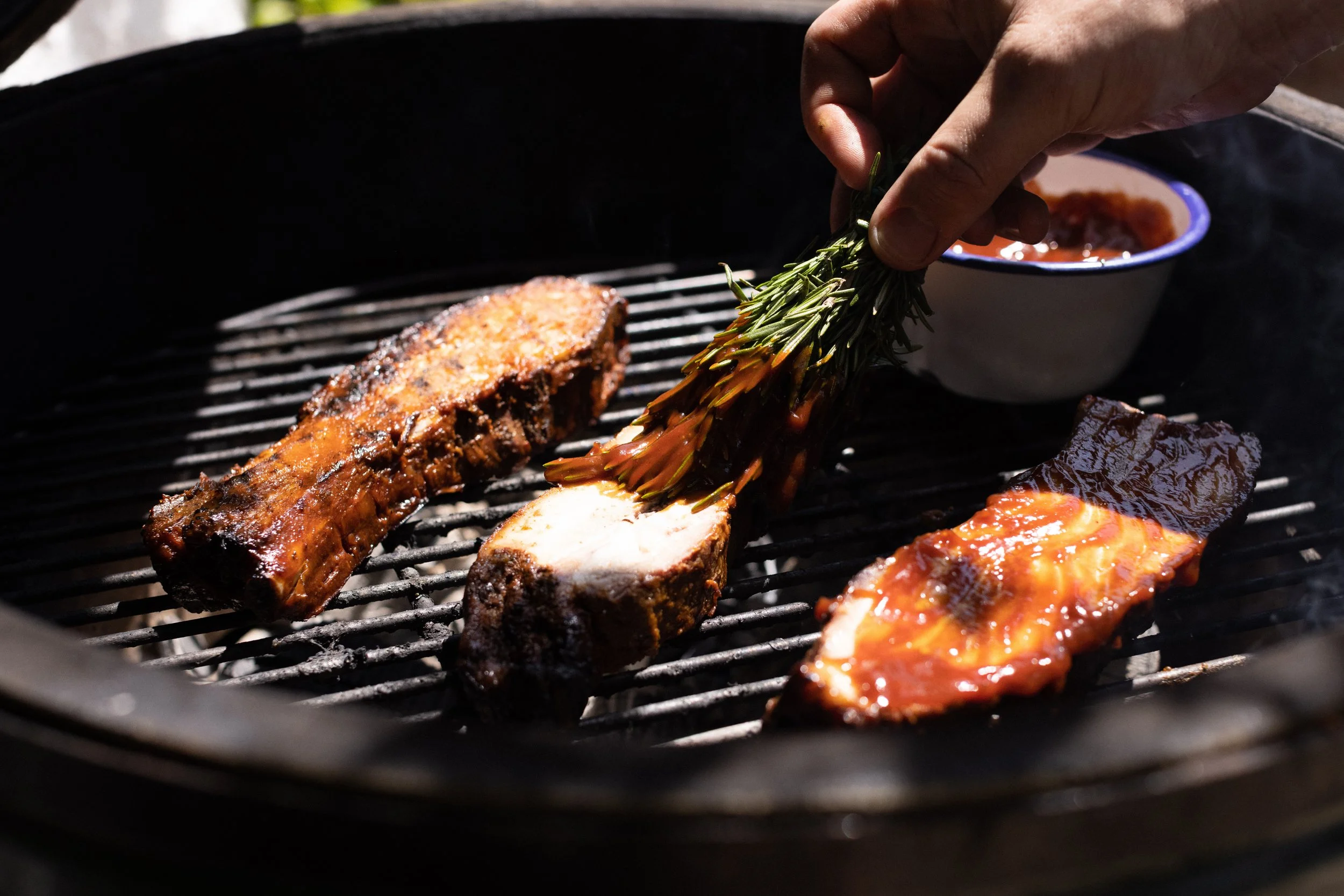 A person placing a sprig of rosemary on a rack of grilling meats over charcoal, with a bowl of sauce in the background.