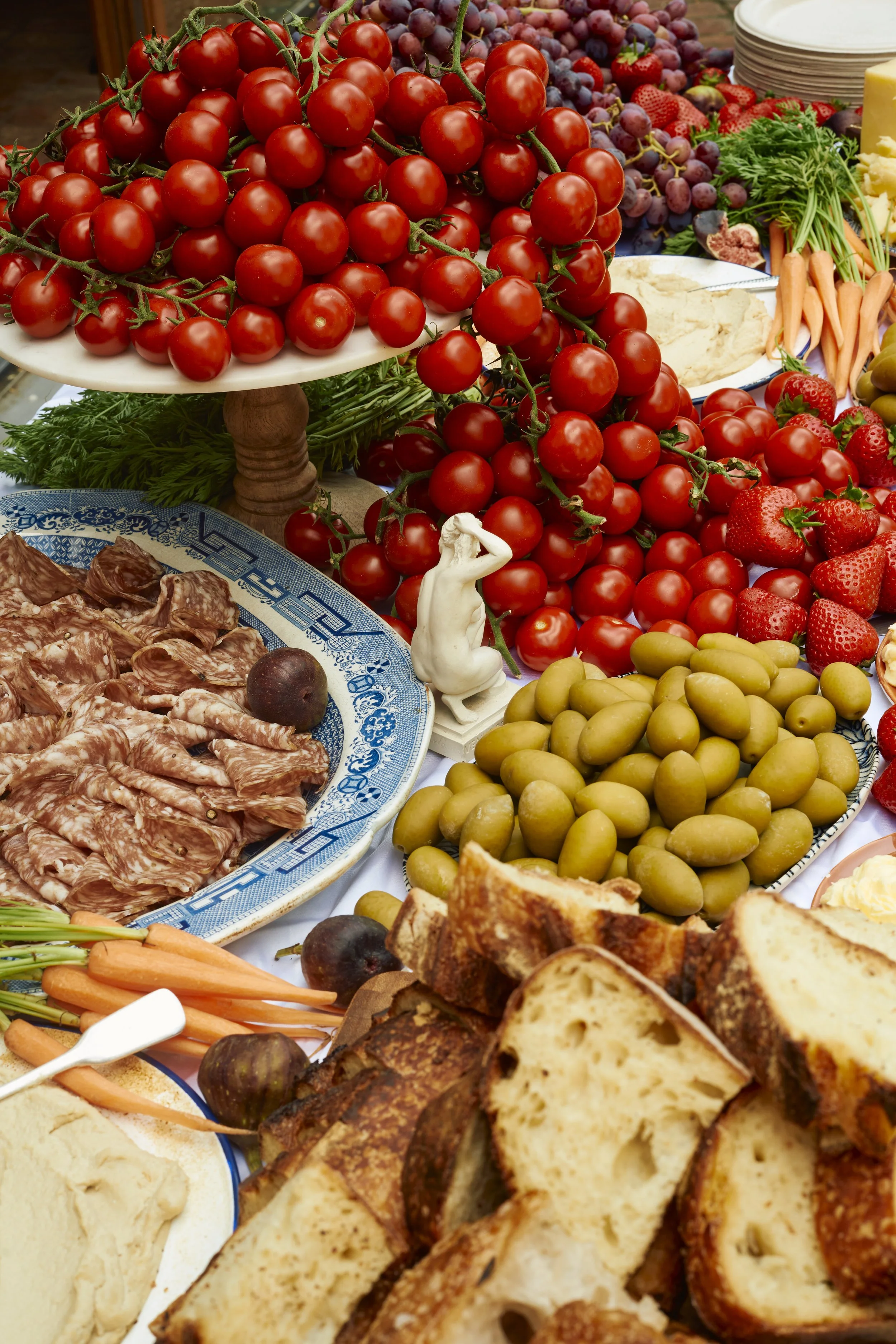 A variety of fresh produce on a table, including stacked cherry tomatoes on a platter, green olives, strawberries, grapes, carrots, and slices of bread, with a small decorative figurine among the items.