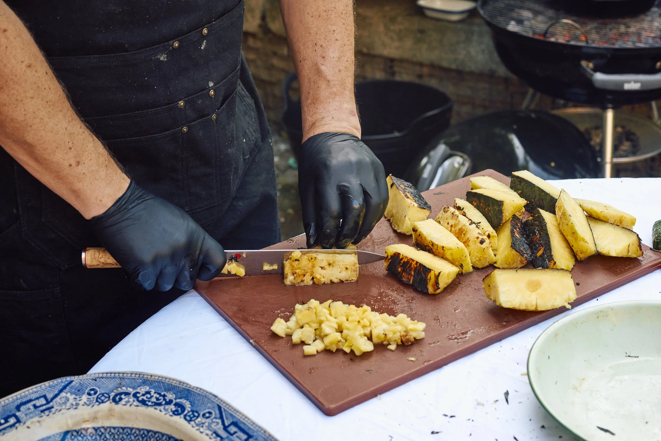 A person wearing black gloves is cutting a large pineapple on a brown cutting board. The pineapple chunks are to the side, with some pieces already sliced. The scene is outdoors with a grill and other objects in the background.