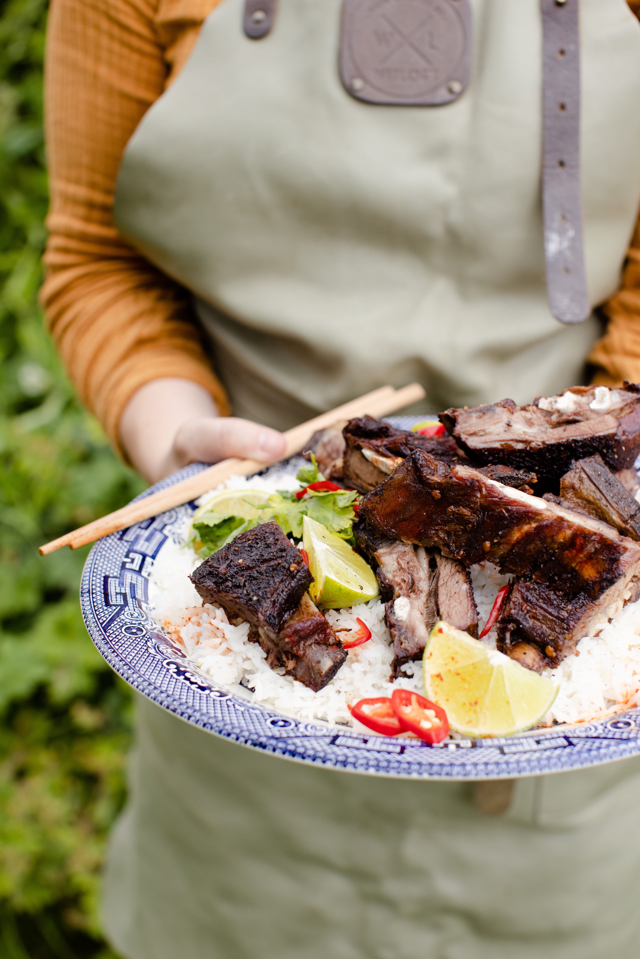 Person holding a blue and white plate with barbequed ribs, rice, lime wedges, and sliced red chili peppers, outdoors.