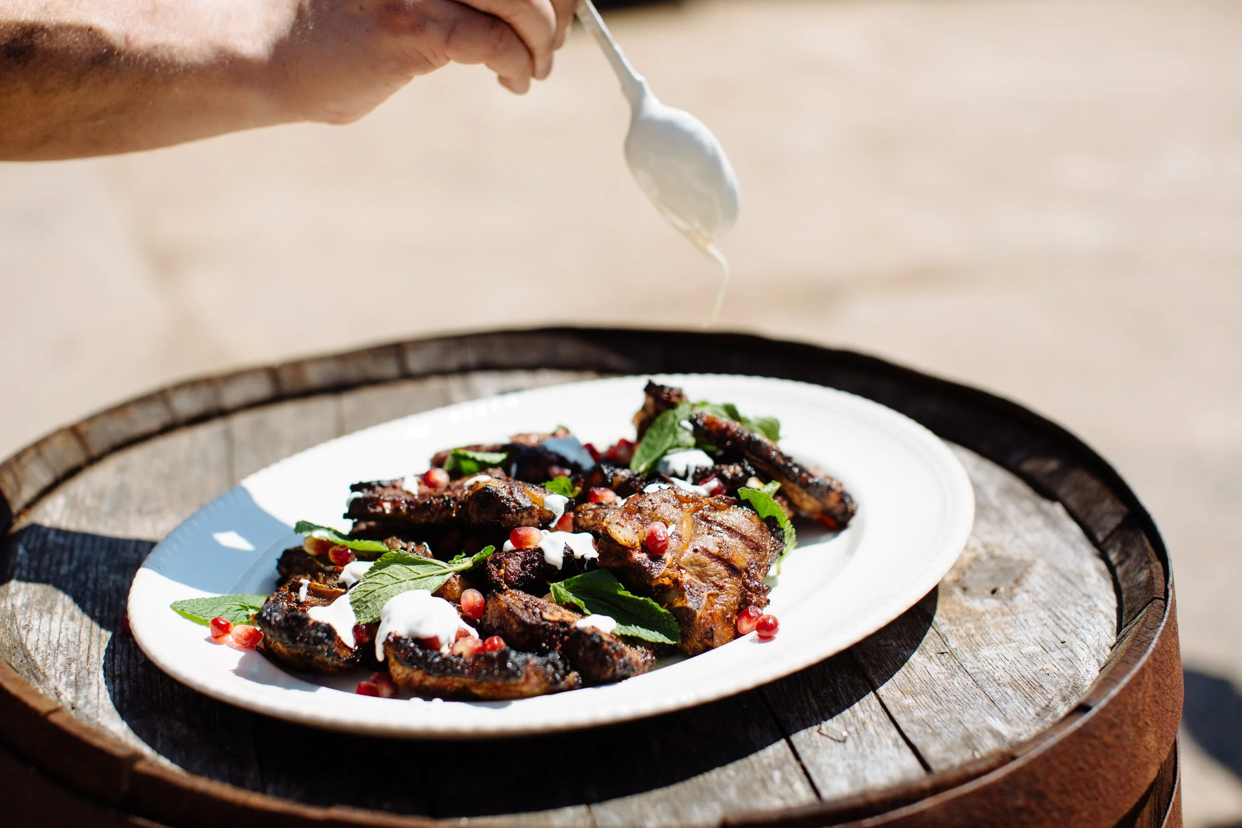 A person is sprinkling sauce over grilled meat and vegetables on a white plate, garnished with fresh herbs and pomegranate seeds, placed on a wooden barrel outdoors.