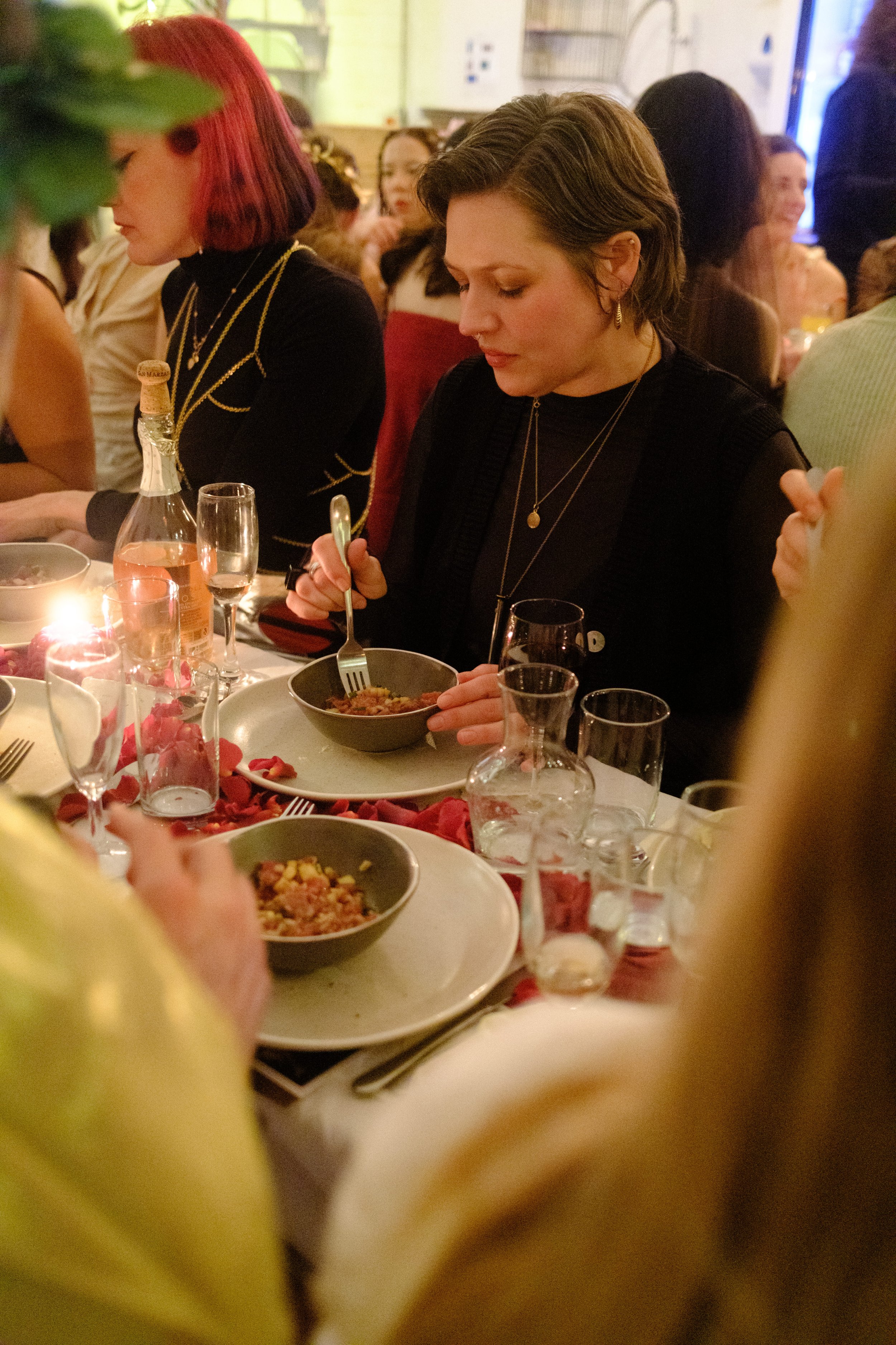 Woman with short brown hair eating at a dinner table with several people, decorated with red petals, candles, and glasses of drinks.