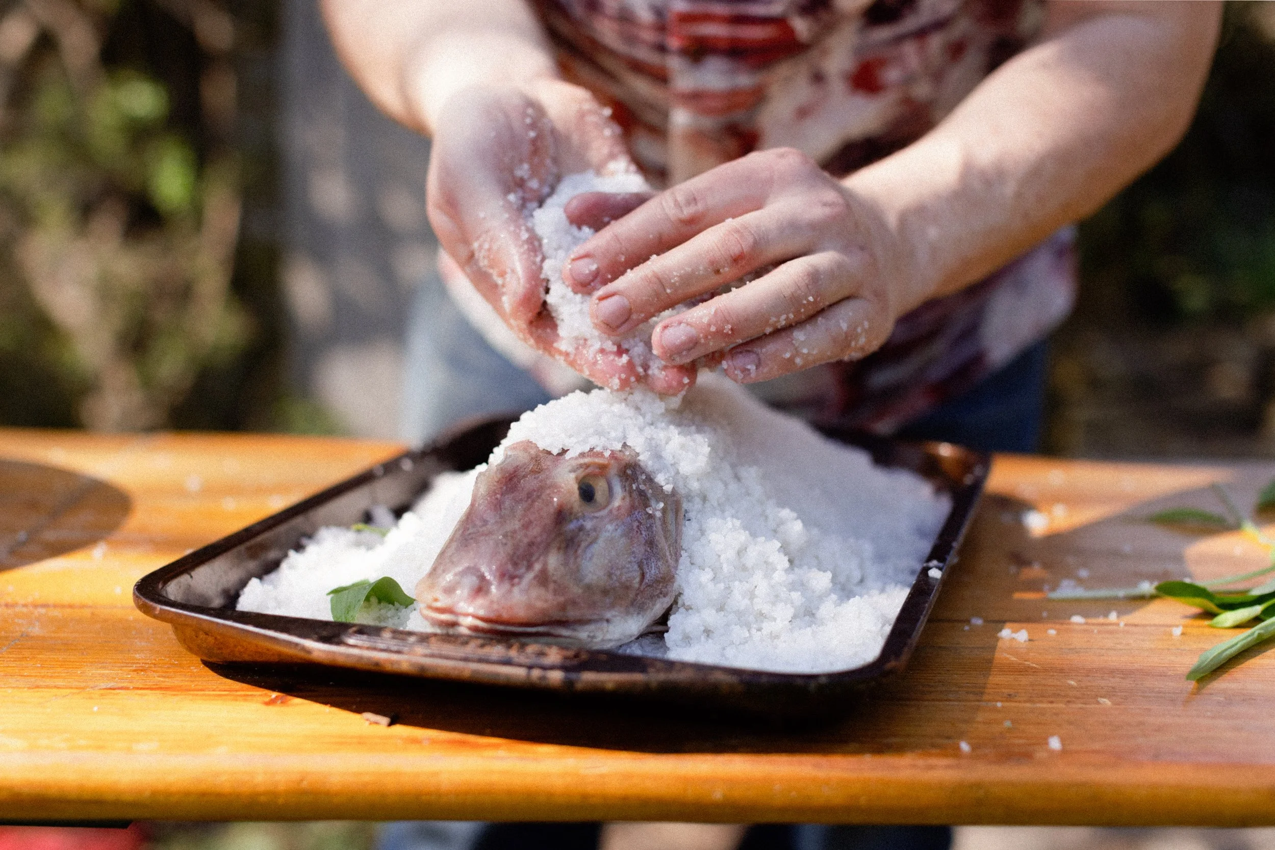 Person seasoning a fish with salt on a black tray outdoors.
