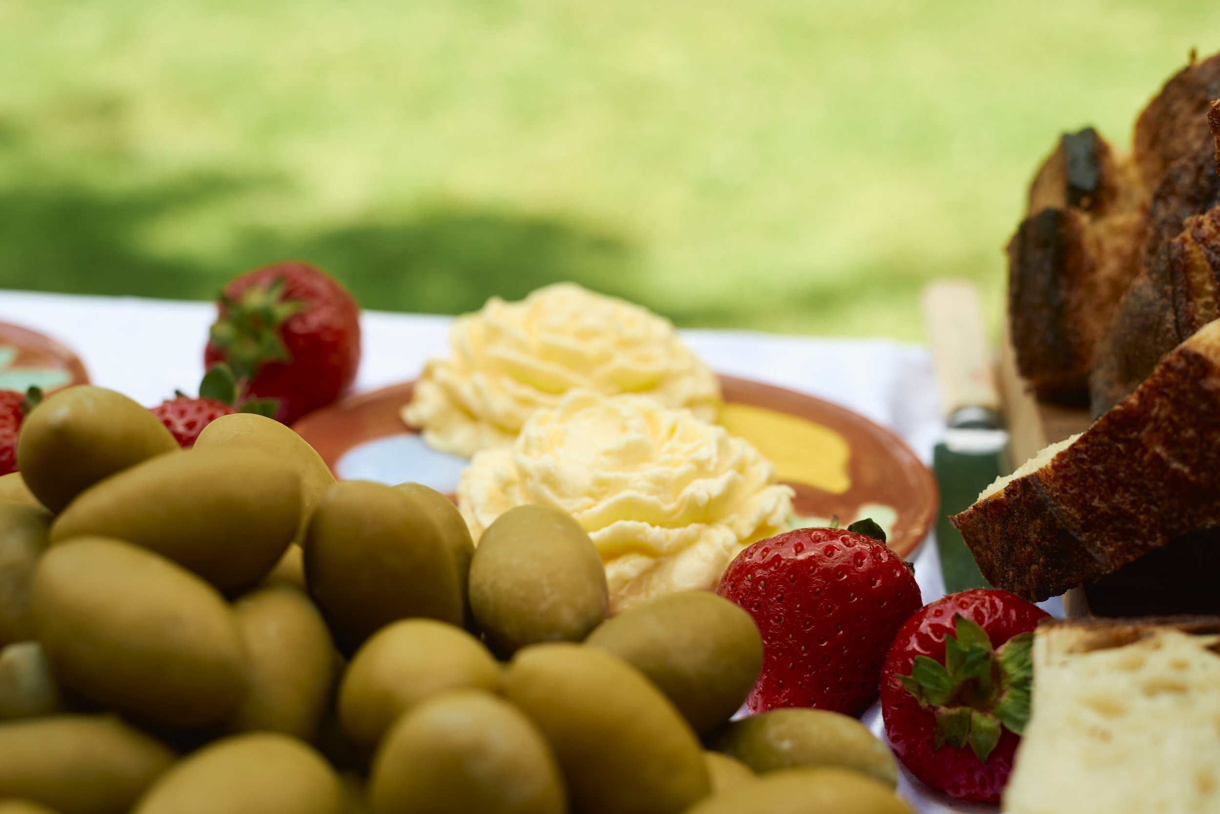 Close-up of a breakfast spread with green olives, strawberries, scrambled eggs, toast, and butter on a plate, set outdoors with blurred green background.