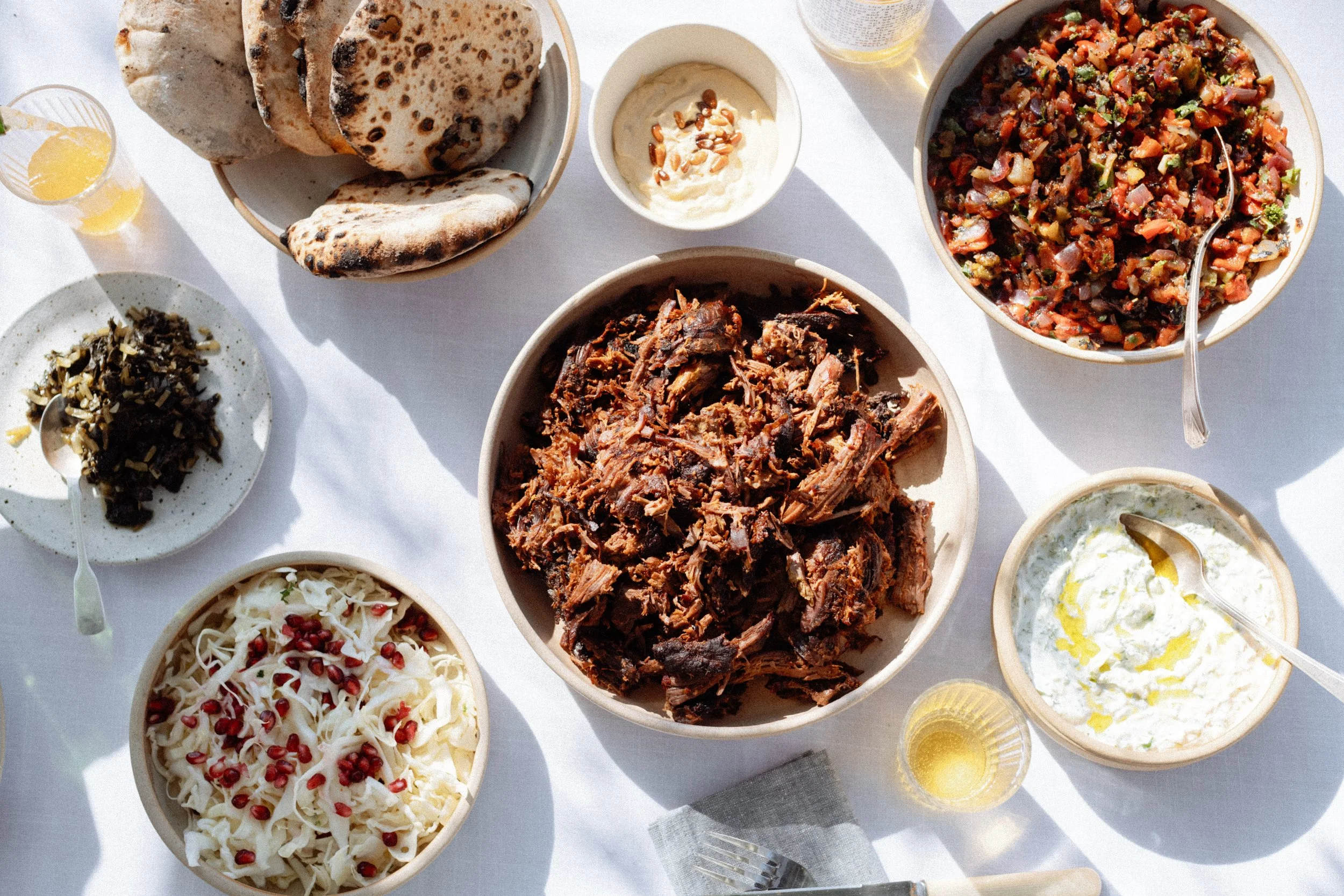 A table set with various Middle Eastern dishes including flatbread, shredded meat, pomegranate seeds with cheese, chopped vegetables, yogurt with herbs and oil, a bowl of stew, and small bowls of sauces and dips.