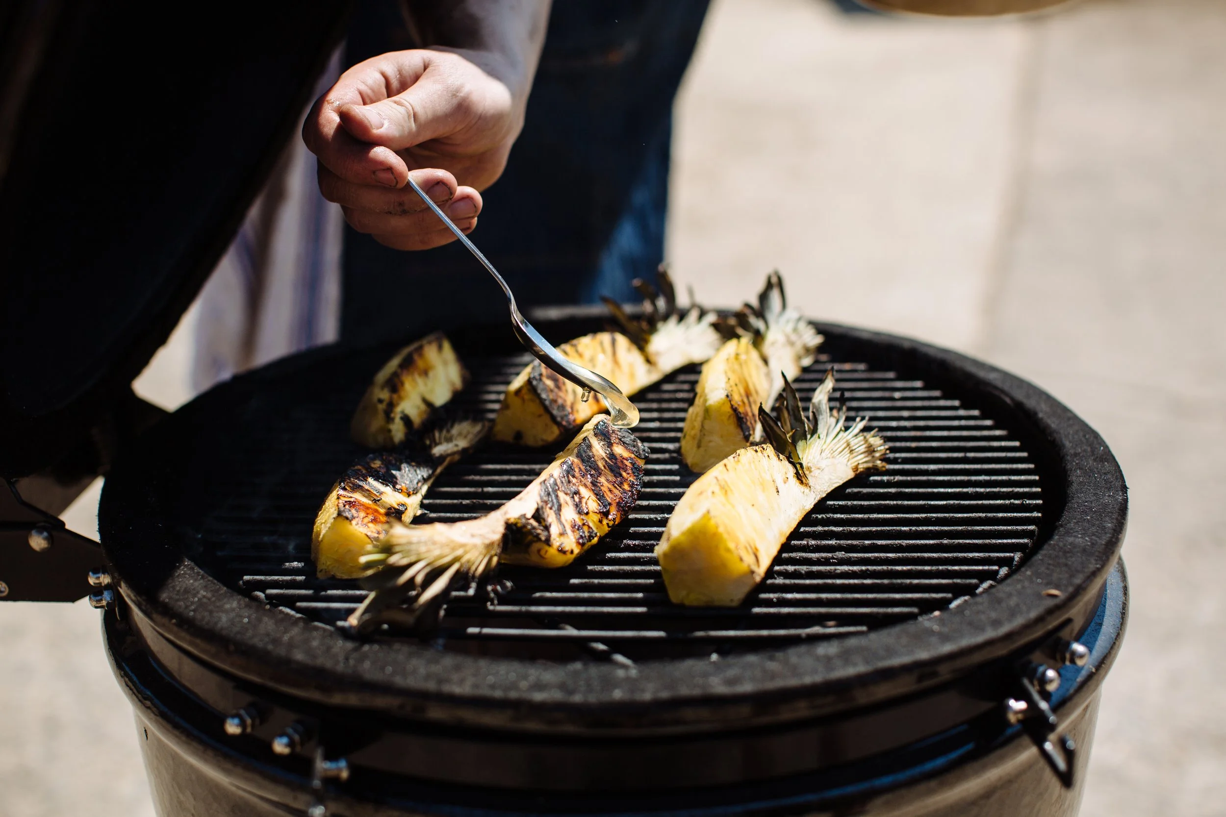 Person grilling pineapple slices on a barbecue grill, using tongs to turn or check the pineapple.