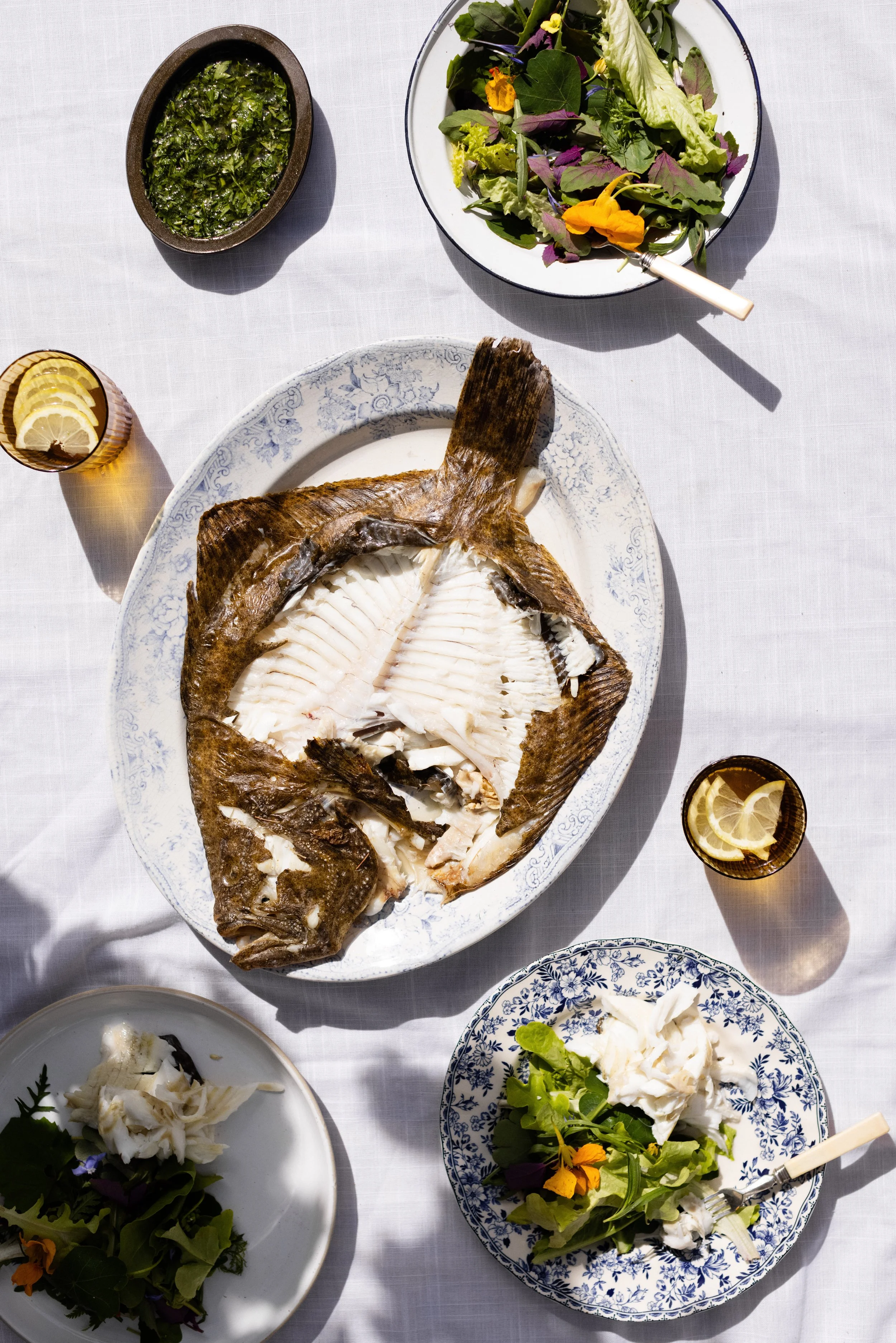 A table set with a large cooked fish on a white plate, two small plates with salad and cheese, two glasses with lemon slices, and bowls of greens, all on a white tablecloth.