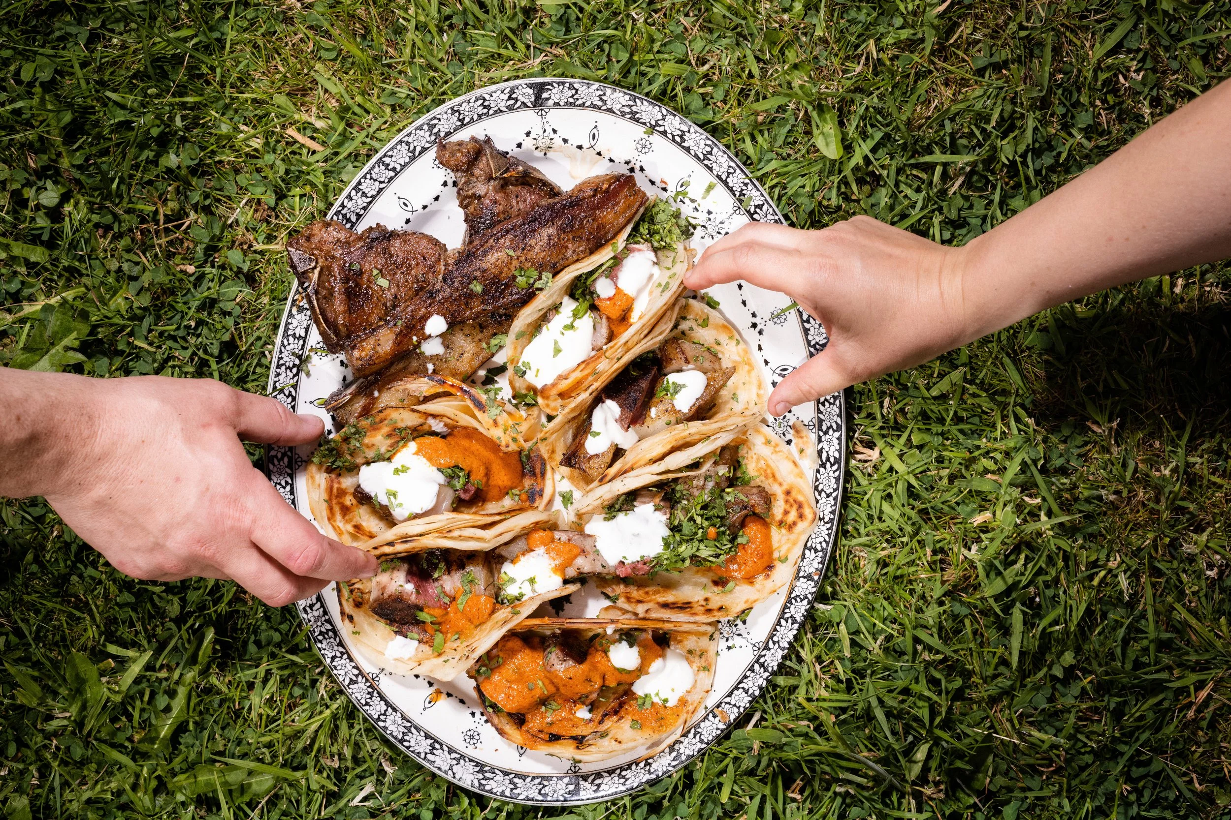Hands reaching for tacos and grilled meat on a decorative oval plate, placed on grass.