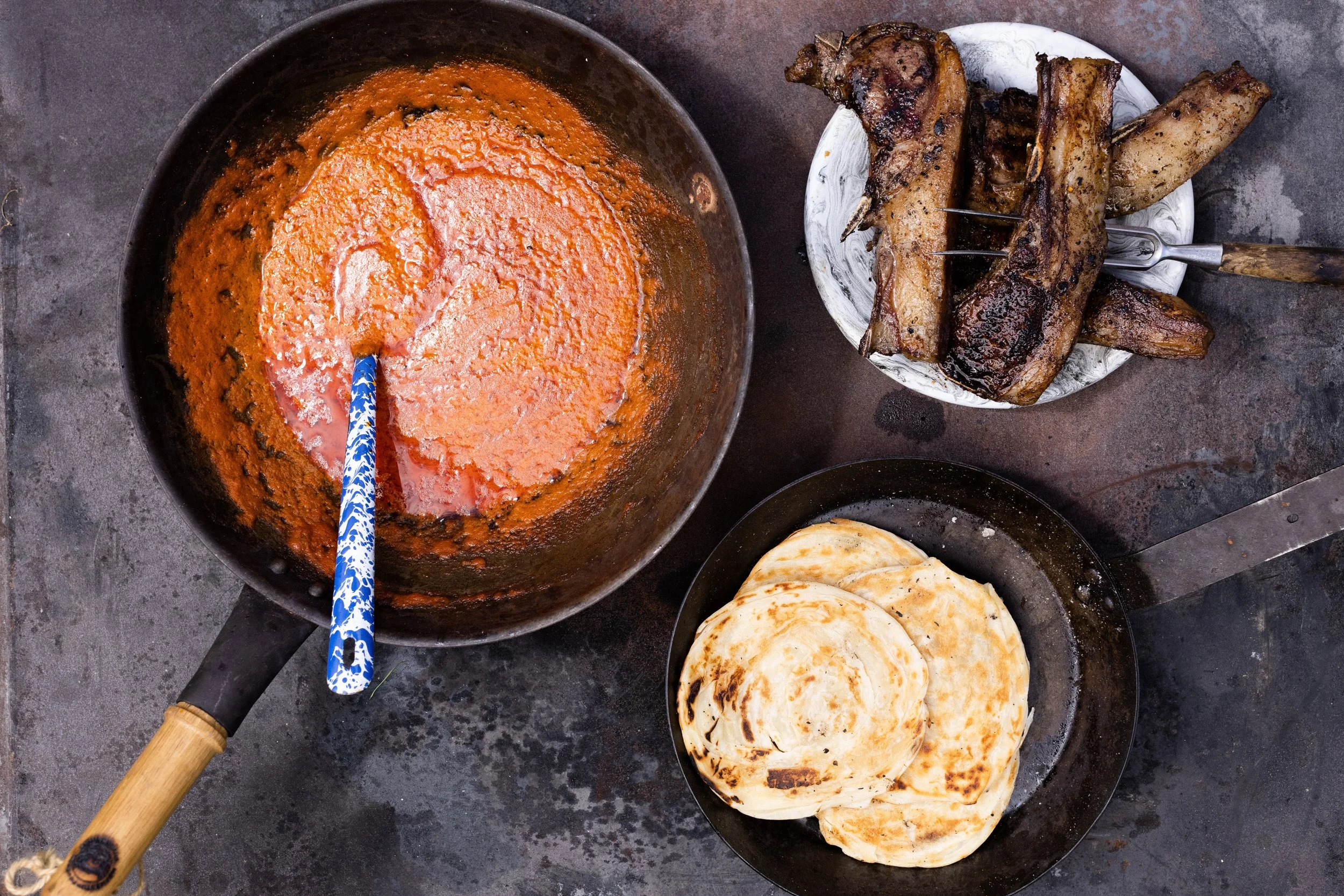 A top-down view of a cooking setup with a pot of red sauce, a plate of cooked ribs, and a pan of tortillas on a dark countertop.
