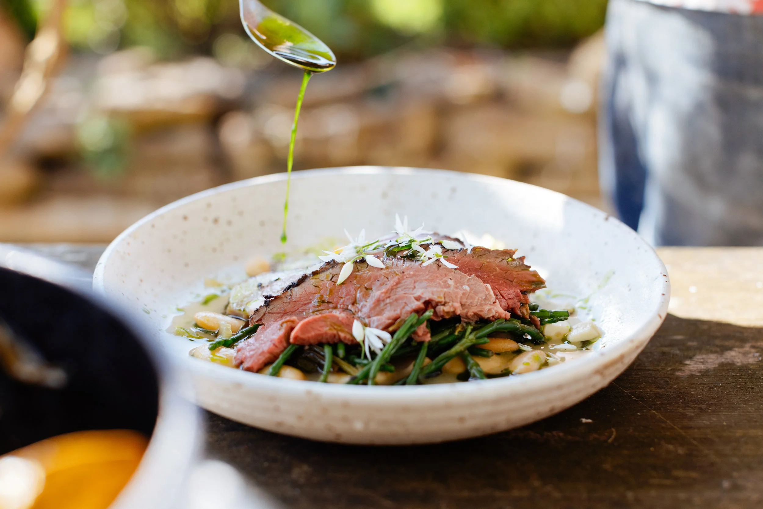 A dish with slices of cooked beef, green vegetables, white beans, and garnished with white edible flowers being drizzled with a green oil or sauce.