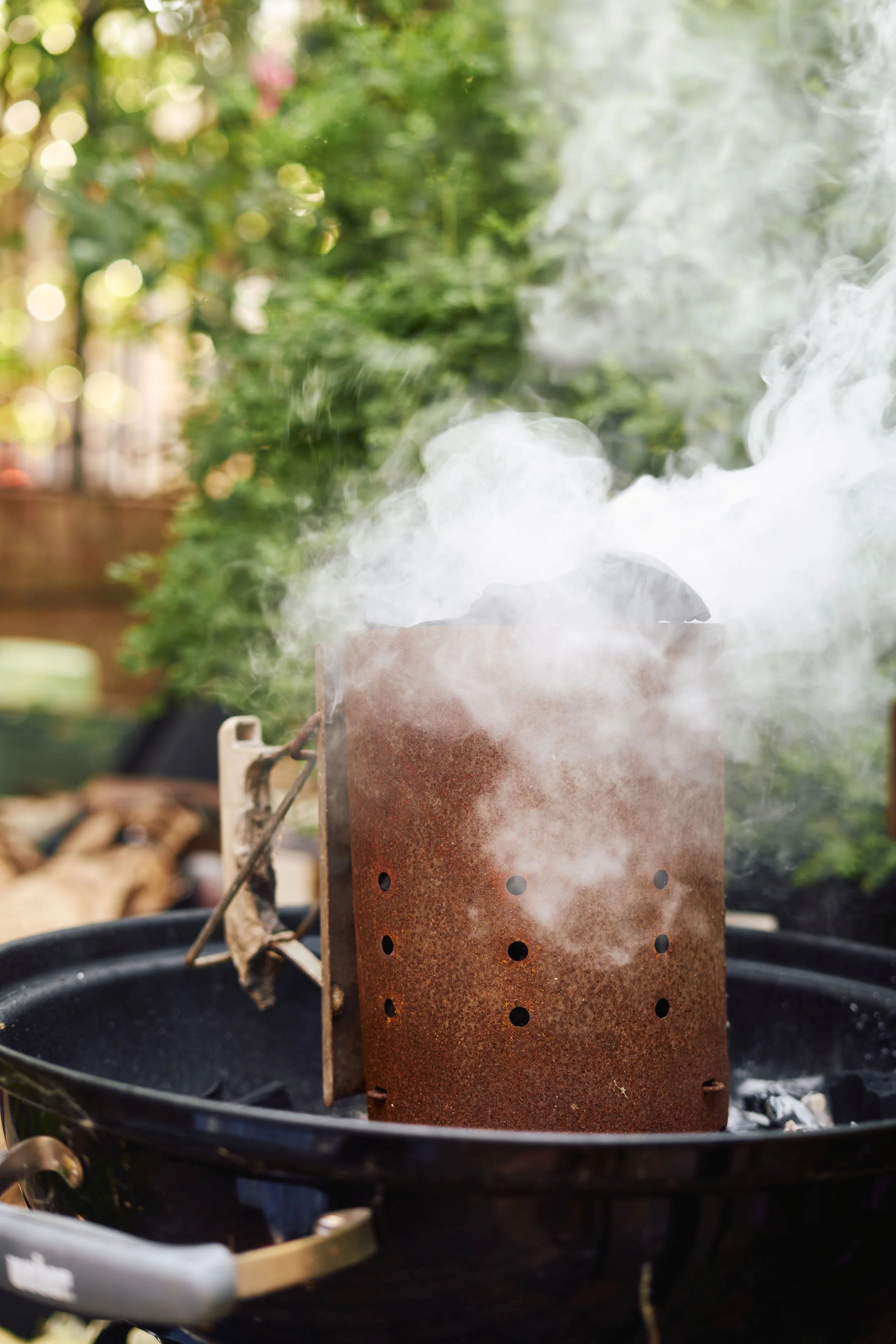 A close-up of a rusted smoke box on a grill, emitting white smoke, with a blurred background of greenery and trees.