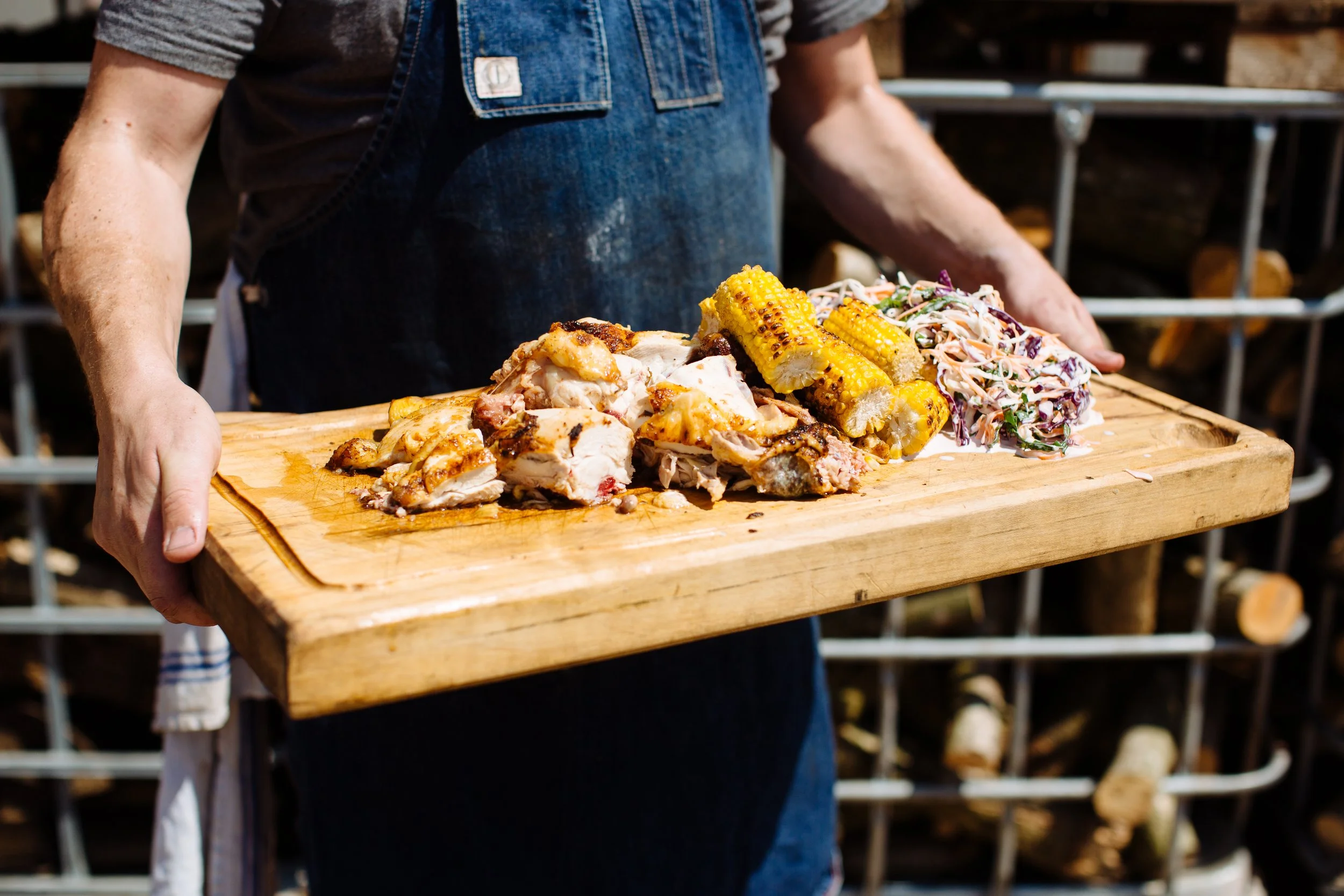 Person holding a wooden platter with grilled chicken, corn on the cob, and coleslaw at an outdoor setting.