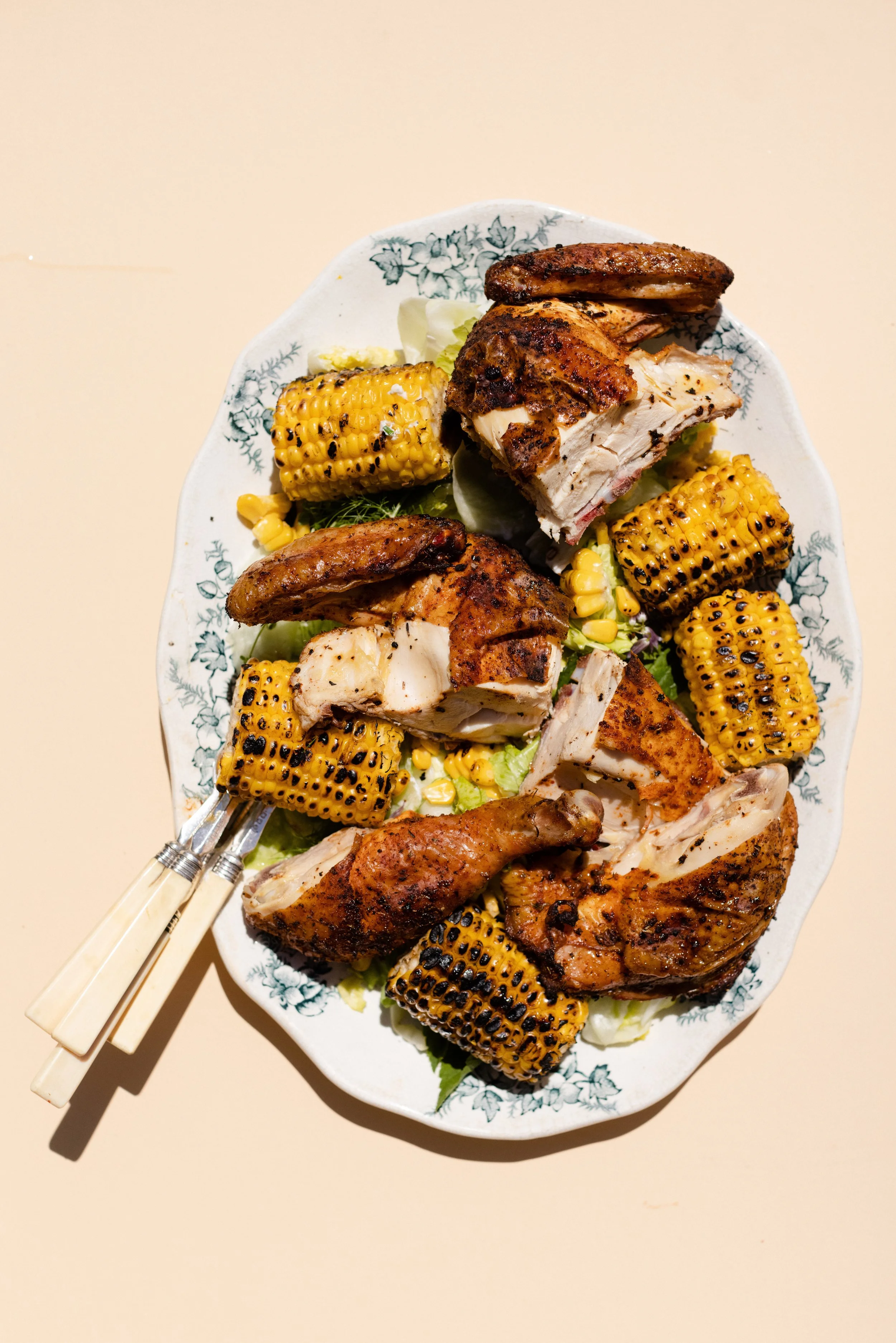 A white plate with a floral pattern holds grilled chicken pieces, grilled corn on the cob, and lettuce leaves, with some chopsticks resting on the side.