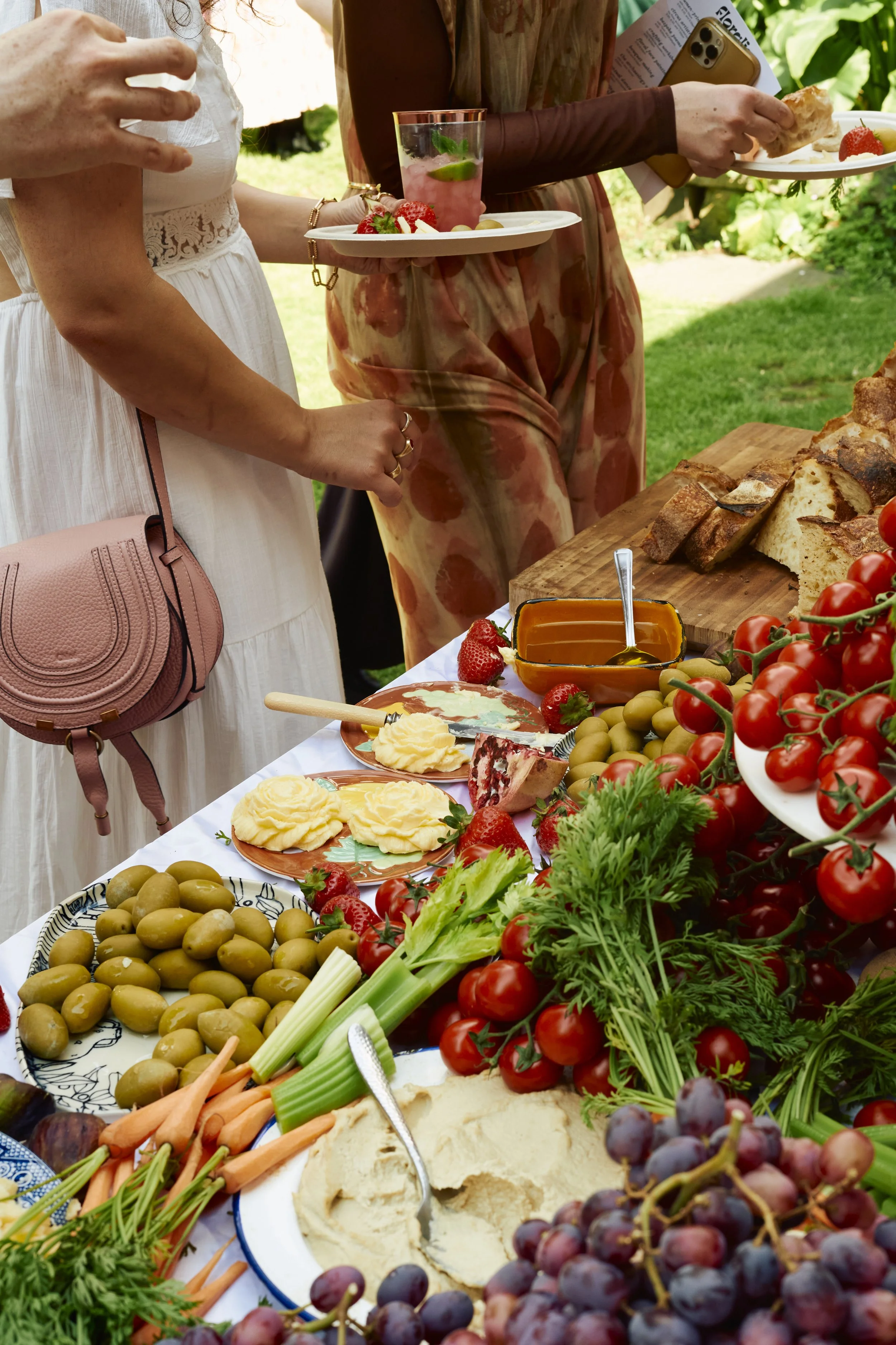A table filled with fresh vegetables, bread, cheese, grapes, strawberries, and dips at an outdoor gathering. People are serving themselves, holding plates with food, in a lush garden setting.
