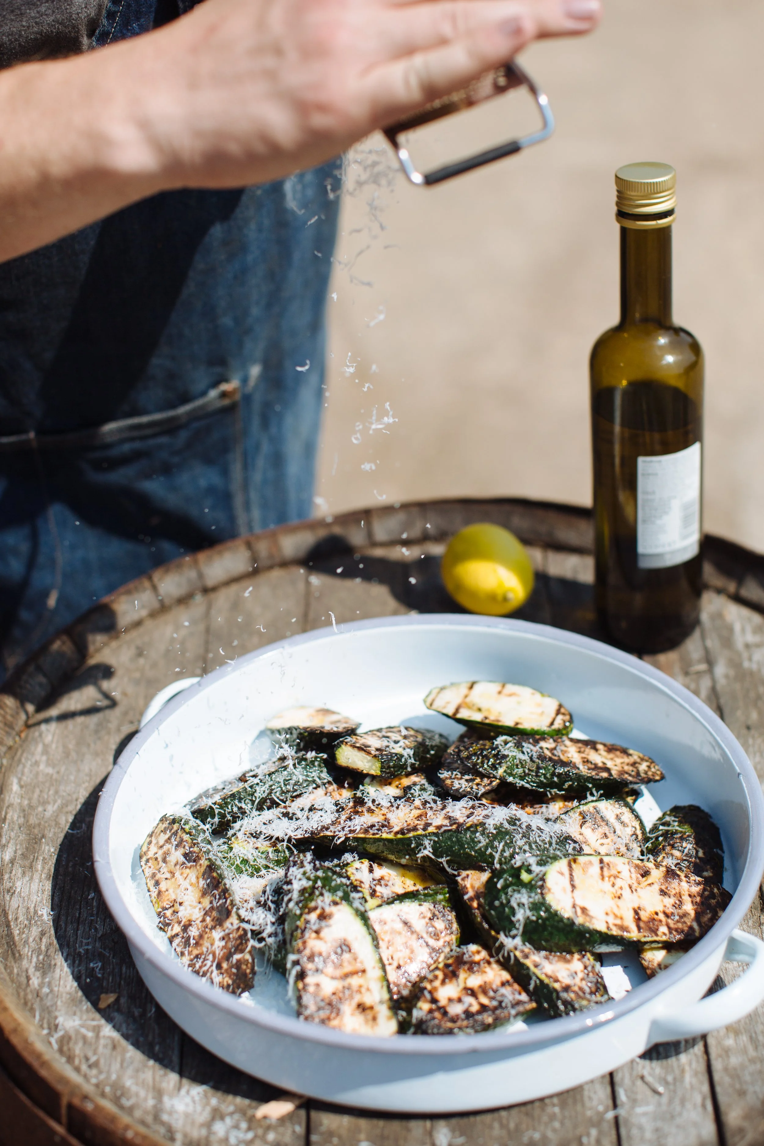 Fried zucchini slices in a white dish with a person sprinkling seasoning or flour on top, on a round wooden surface outdoors, with a lemon and a bottle of oil in the background.