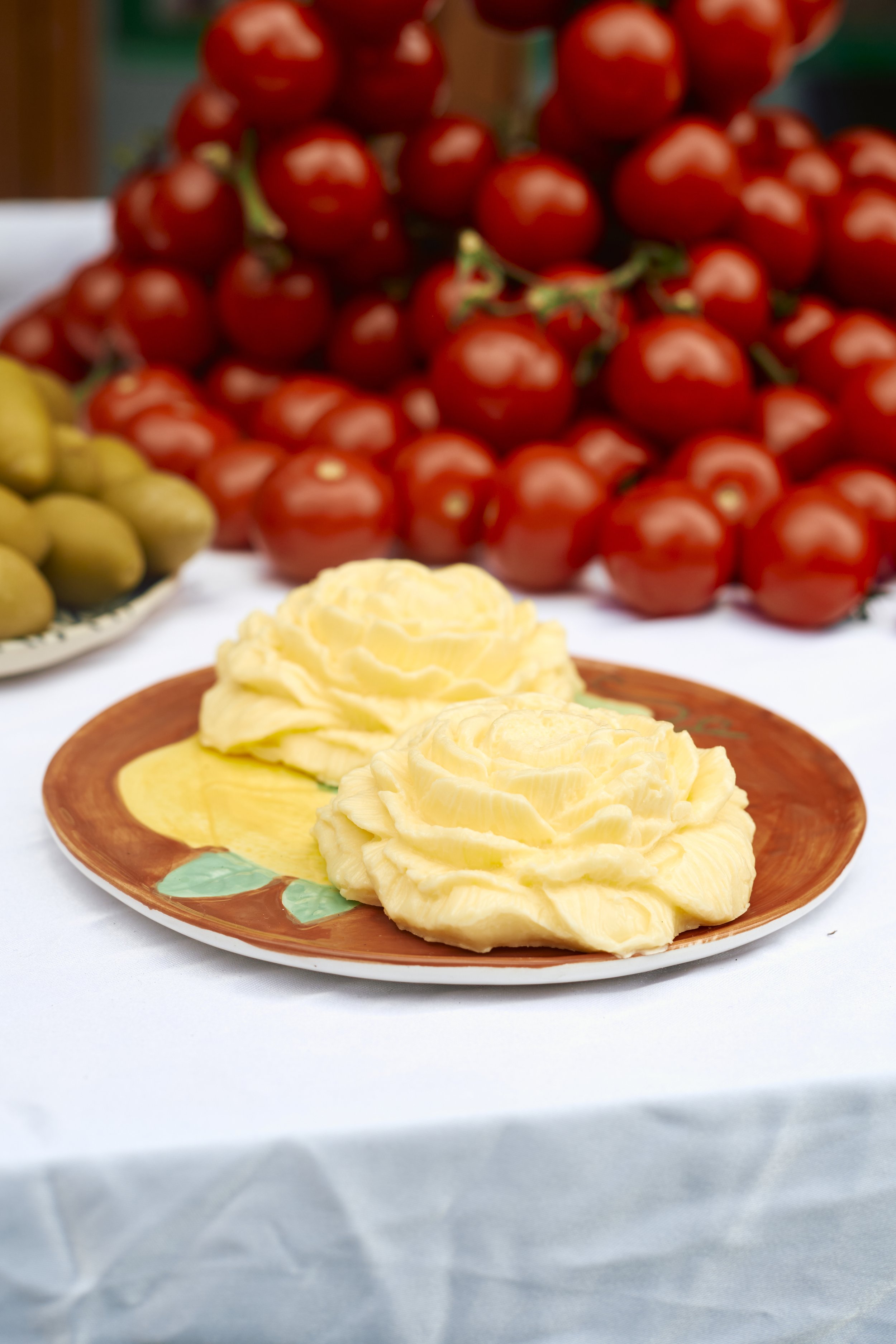 Two dollops of yellow butter on a floral-patterned ceramic plate, with a background of cherry tomatoes and green olives on display.