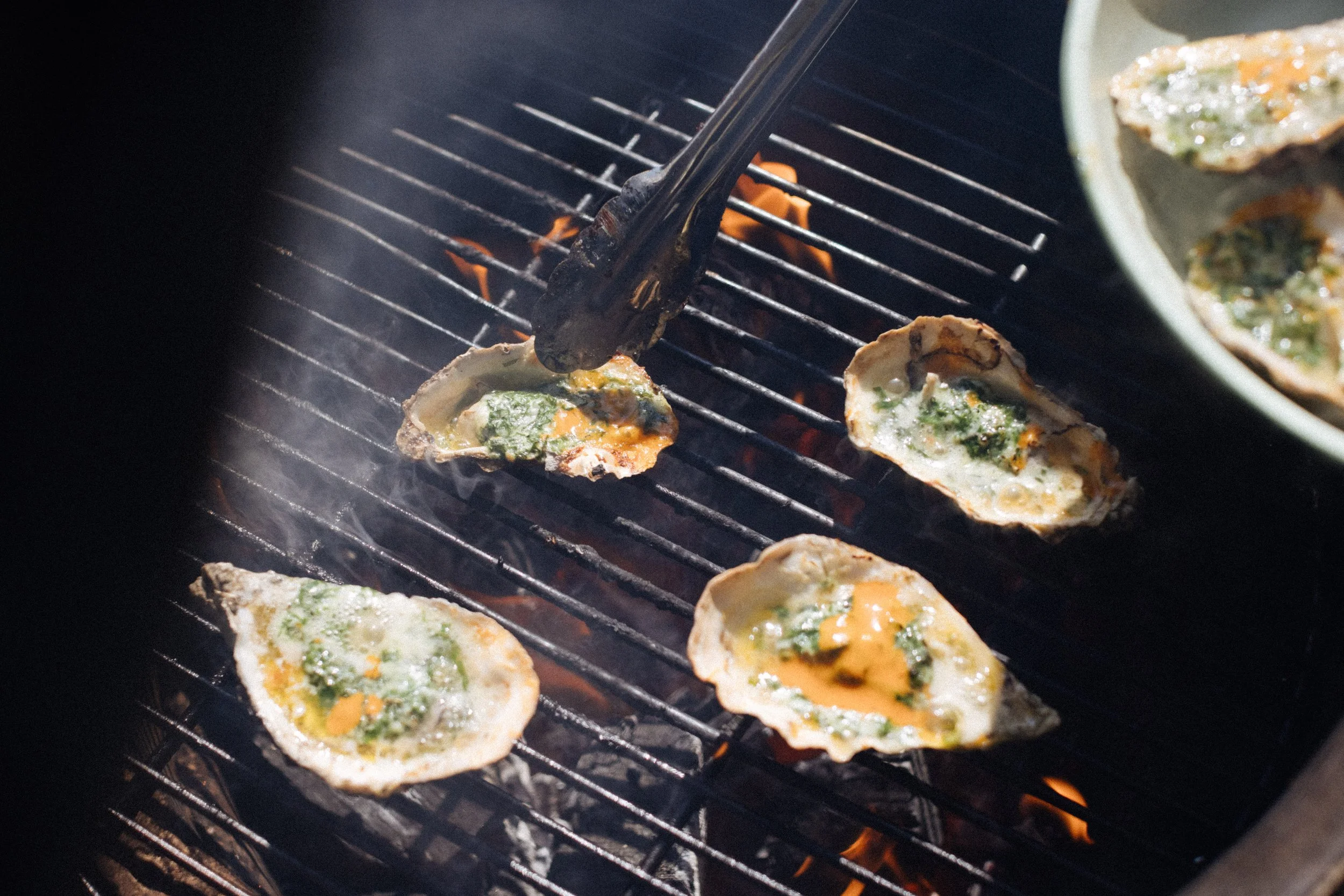Oysters being grilled on a barbecue grill, with some being handled by tongs.