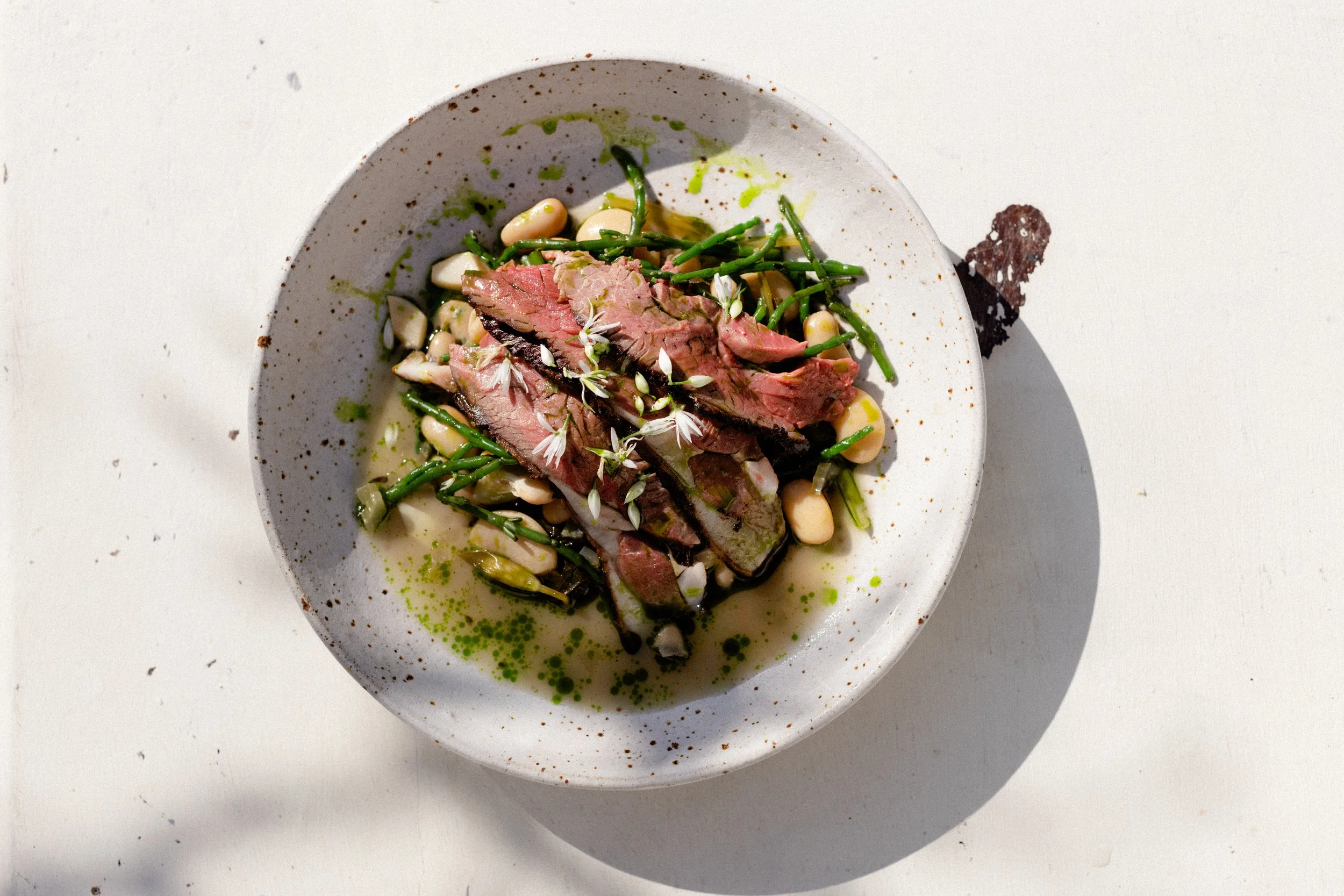 A dish with sliced medium-rare beef, green beans, white beans, and a drizzle of green sauce, garnished with small white edible flowers in a speckled ceramic bowl on a white surface.