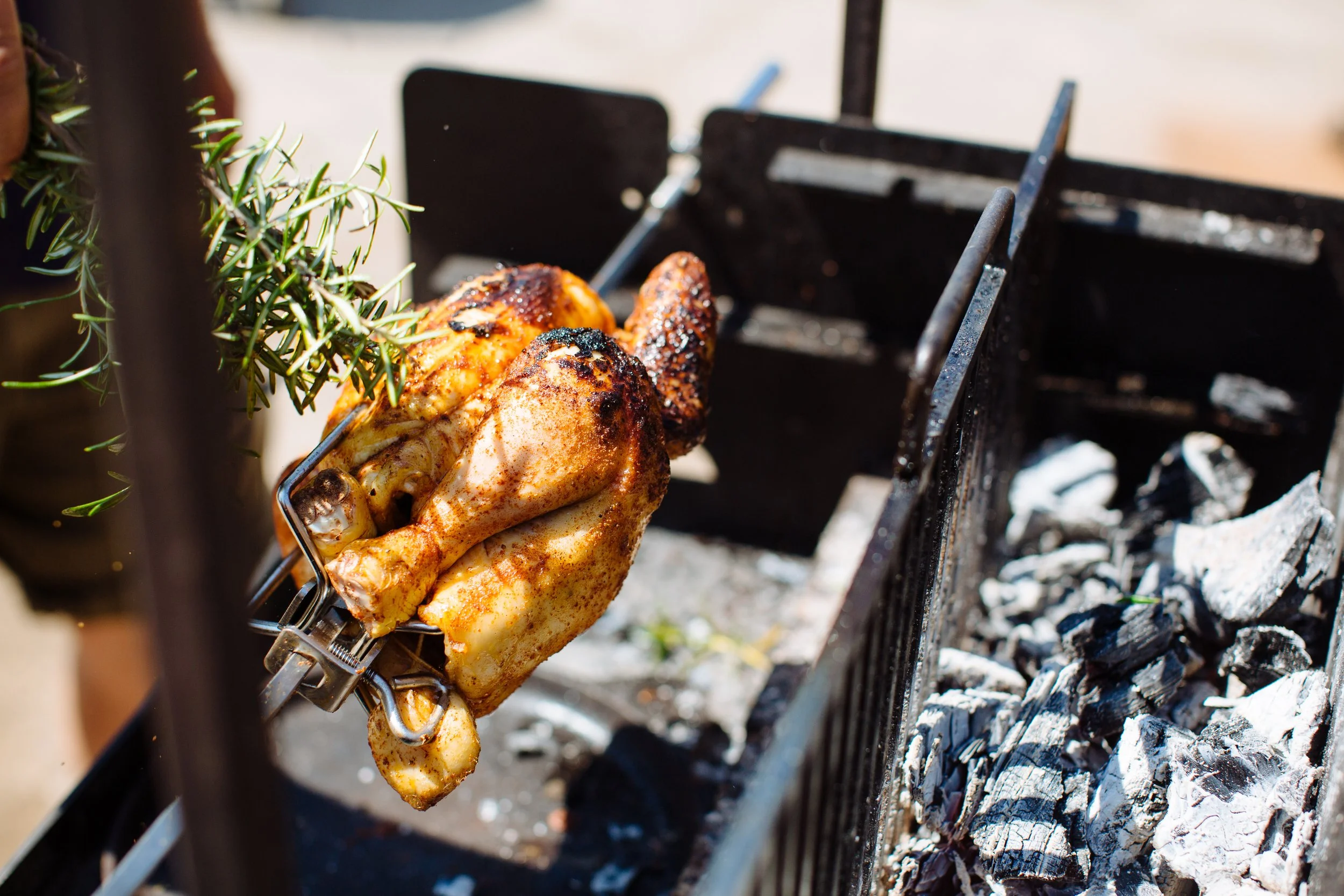 Chicken being roasted on a rotisserie over charcoal with a sprig of fresh rosemary nearby.