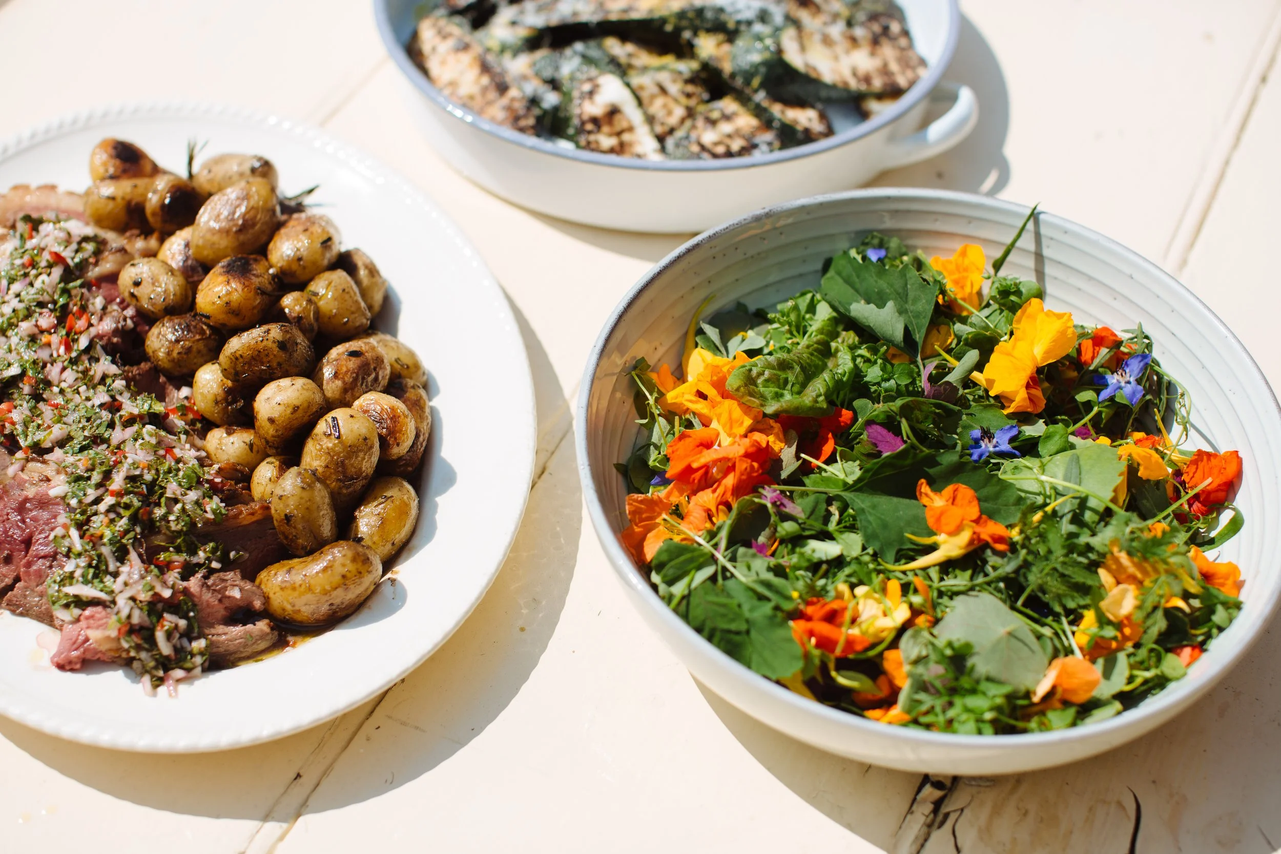 Plate of roasted new potatoes with herbs and slices of rare beef topped with herbs, two bowls of salad with mixed greens and edible flowers, and a bowl of grilled vegetables in the background.