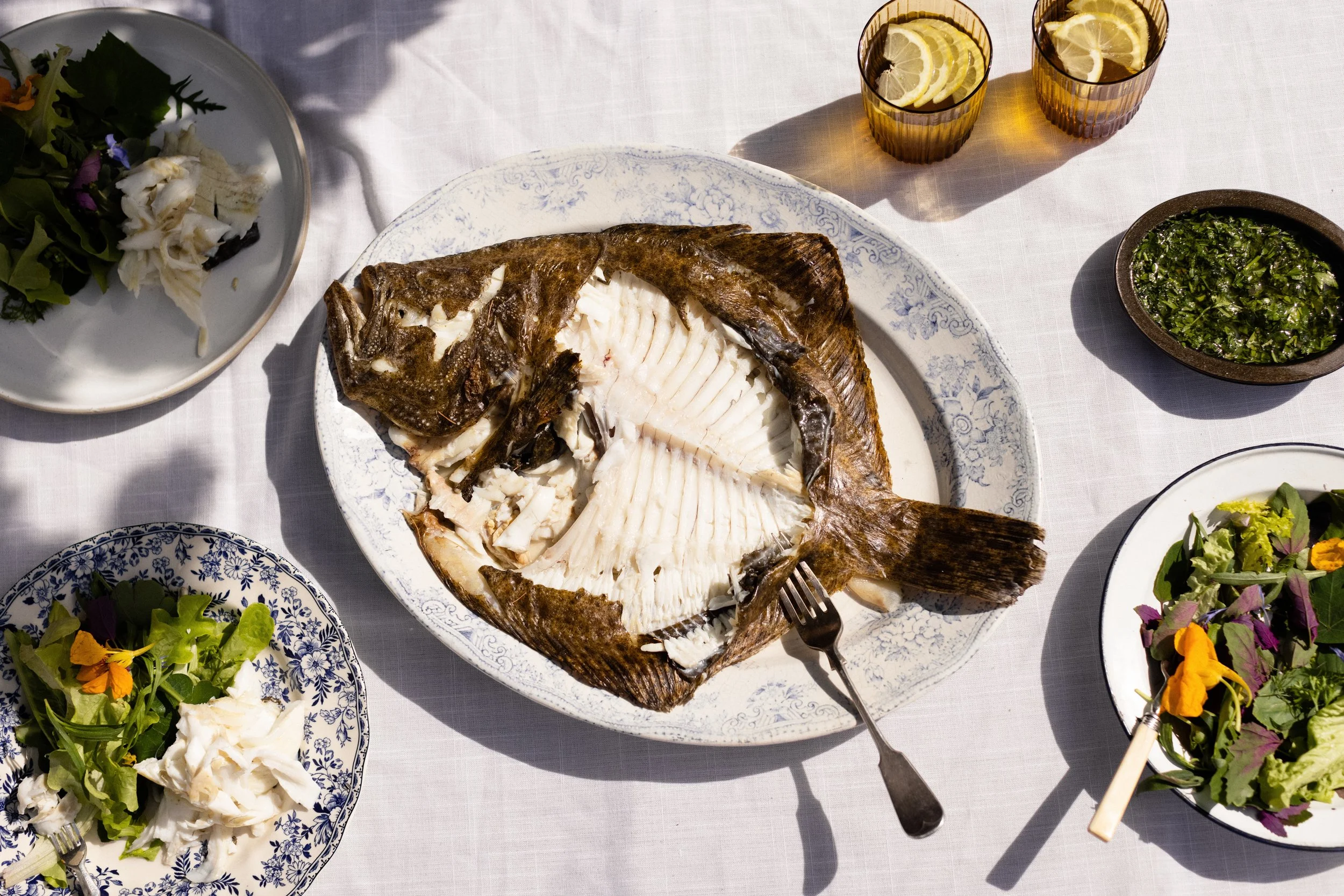 A whole cooked fish on a white platter with a fork, surrounded by plates of salad, and glasses of lemon water on a white tablecloth.