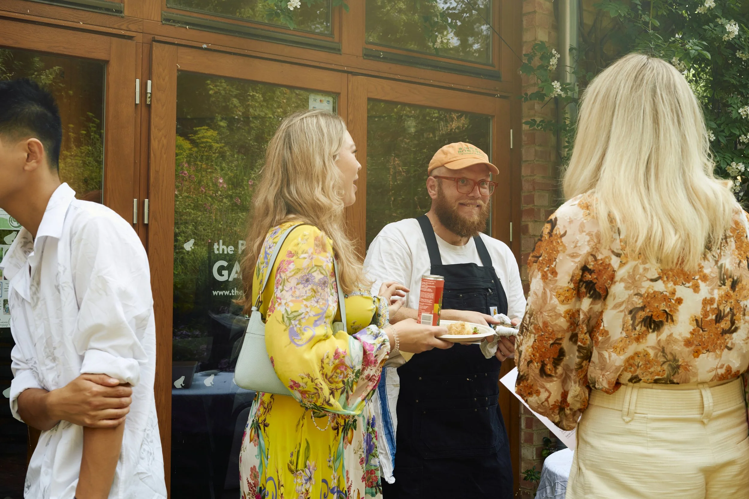 A group of people socializing outside a building, one woman holding a plate of food and a man with a beard and glasses, while another woman faces away from the camera in a floral shirt.